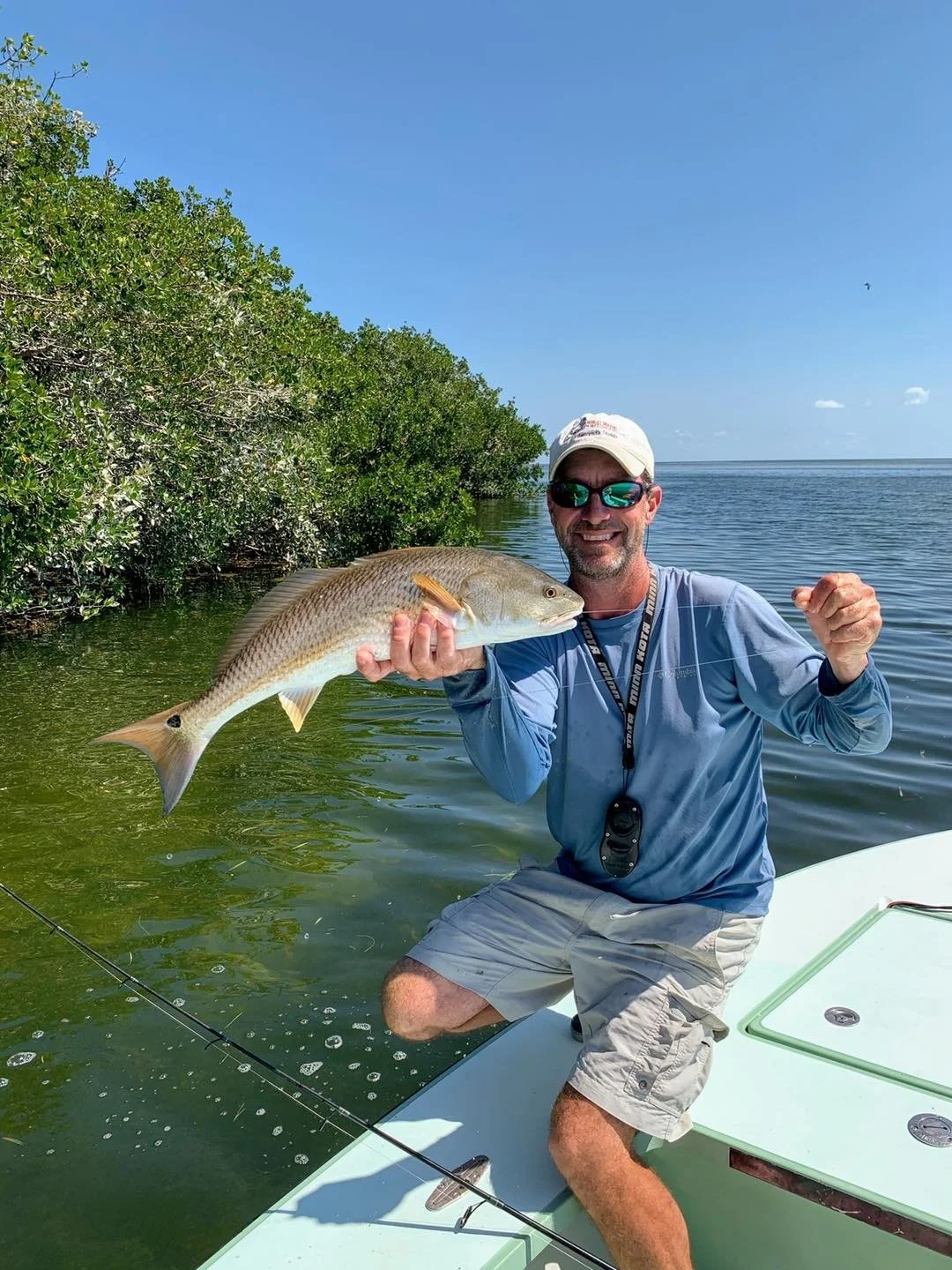 Man holding a large fish on a boat near a mangrove shoreline on a sunny day.