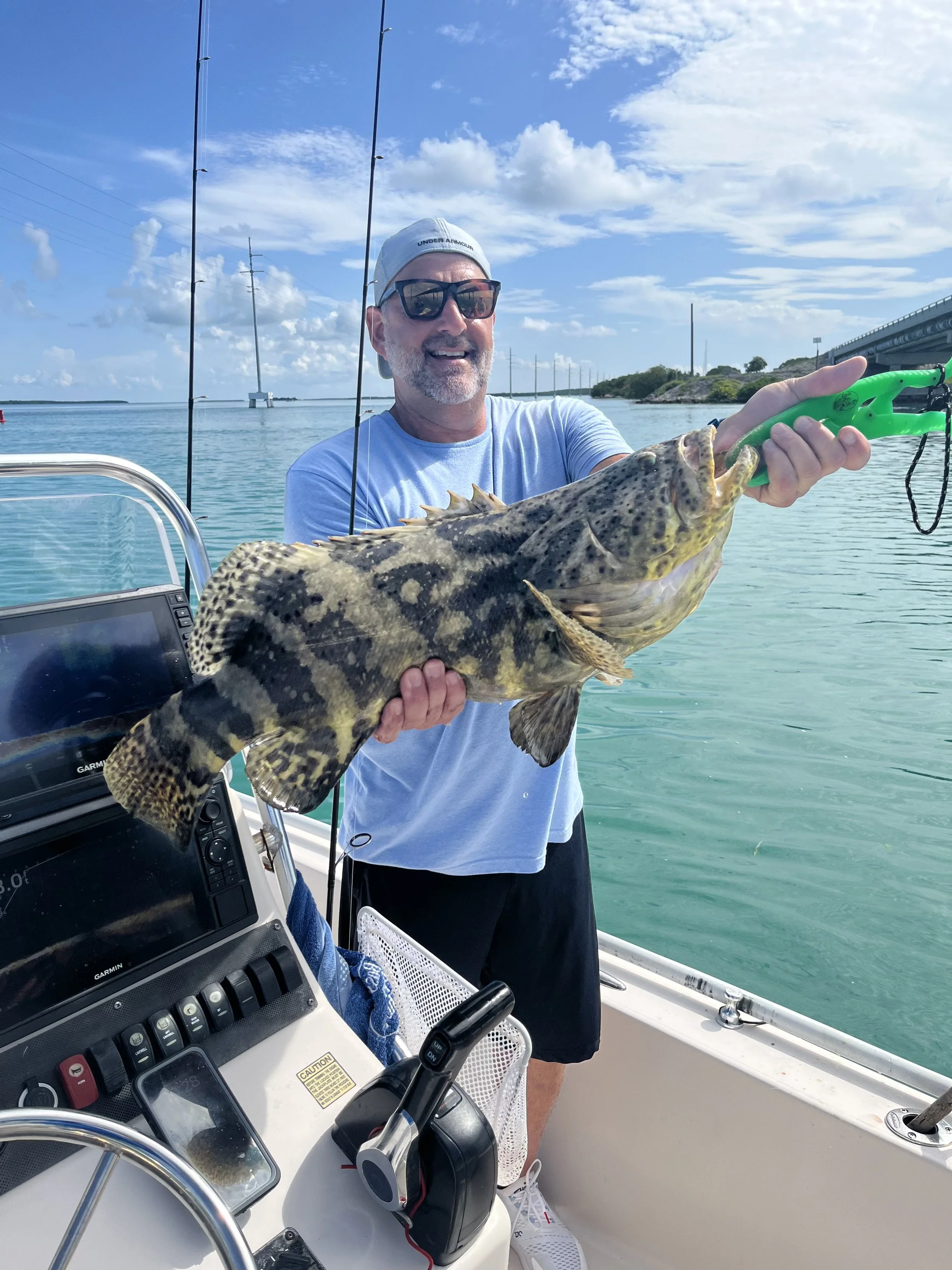 A man wearing sunglasses and a baseball cap happily displays a large fish while on a boat in clear blue water under a partly cloudy sky.