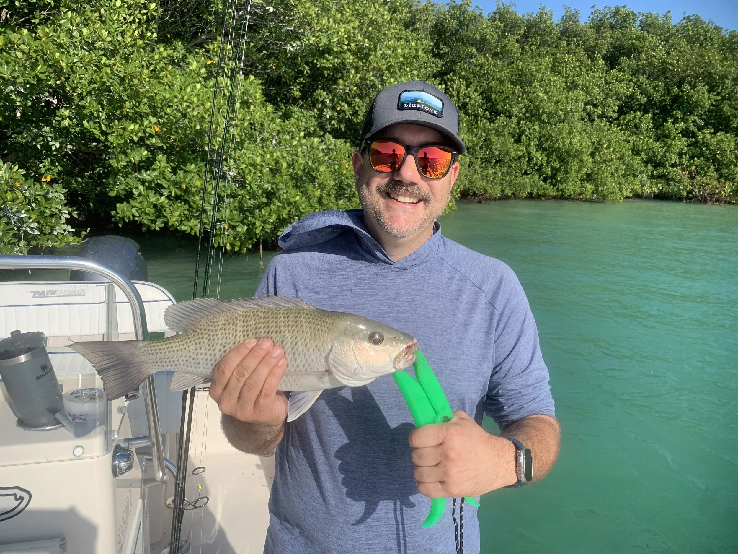 Man with sunglasses and cap holding a fish on a boat in clear water with green mangrove trees in the background.