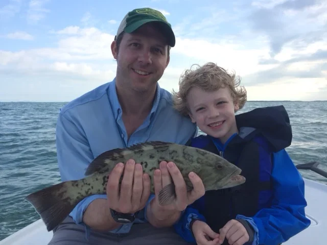 A man and a young boy sitting on a boat holding a fish. They are smiling at the camera with water and a cloudy sky in the background.