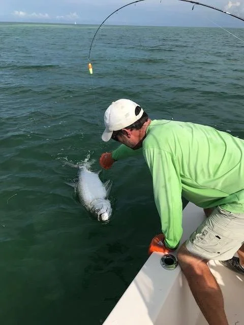 A man in a green shirt and white cap fishing from a boat, holding a fish in the water.