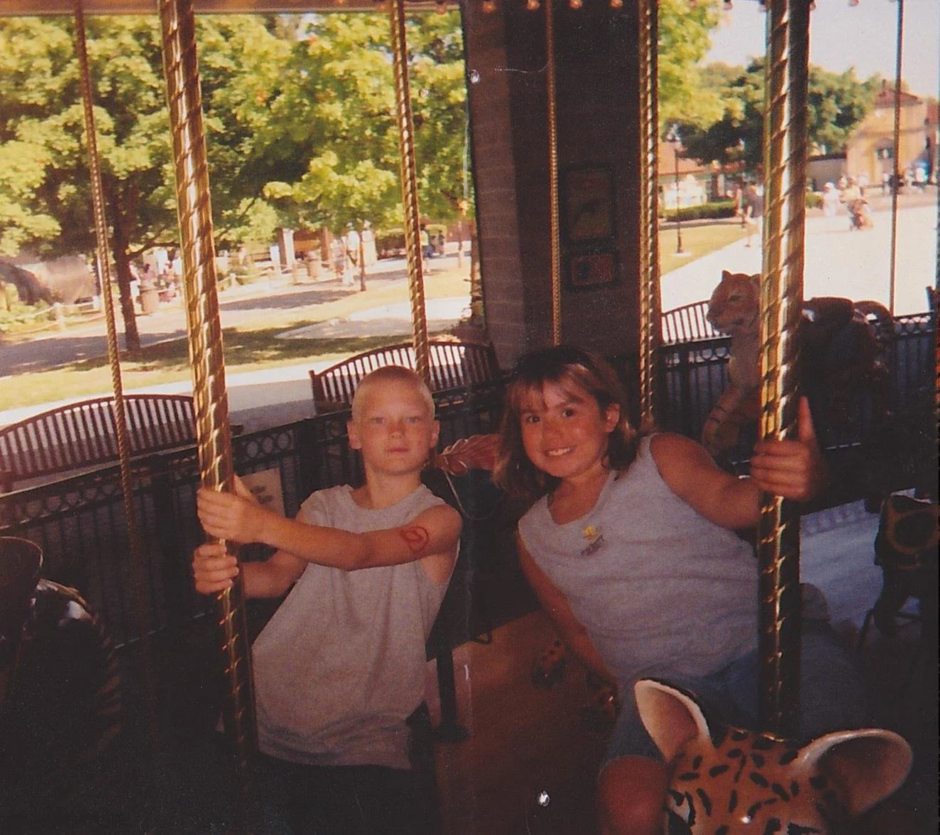 Two children, a boy and a girl, riding a carousel at an amusement park, with the boy on the left holding a carousel pole and the girl smiling and holding another pole, with outside trees and park visible through the carousel window.