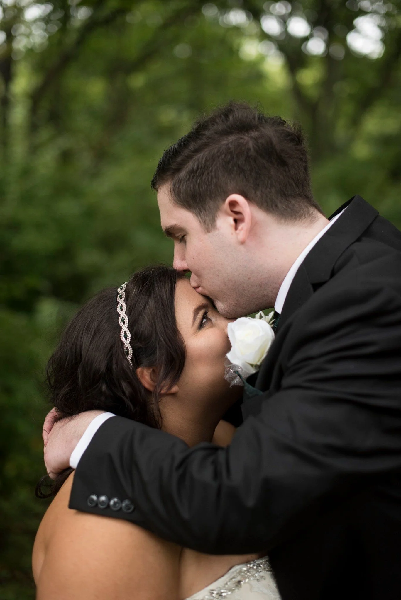 A groom kisses a bride on the forehead outdoors, surrounded by green trees, with the bride holding a white flower lapel.