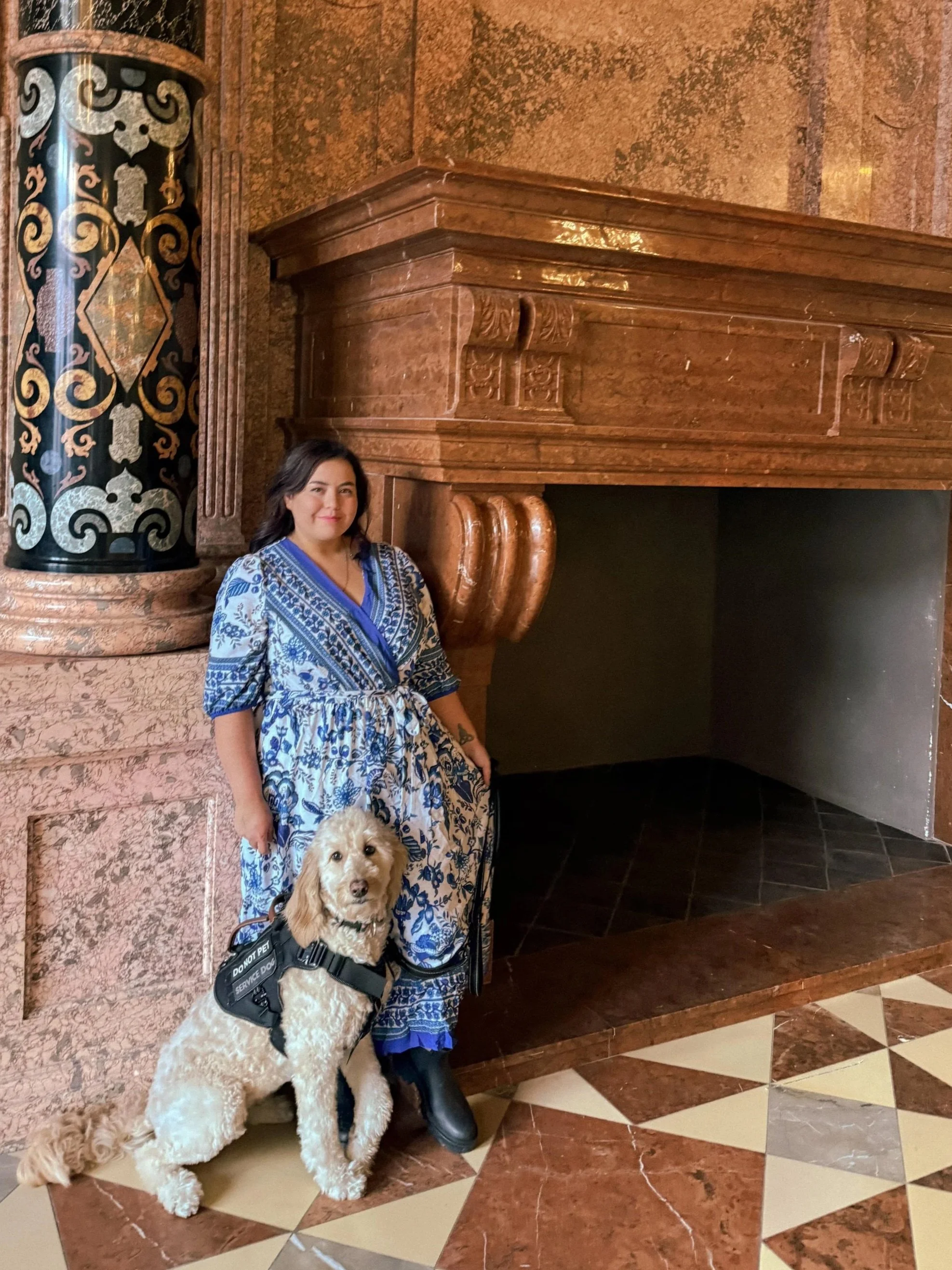 A woman in blue and white patterned dress standing next to a service dog inside a building with ornate wooden and marble decor.