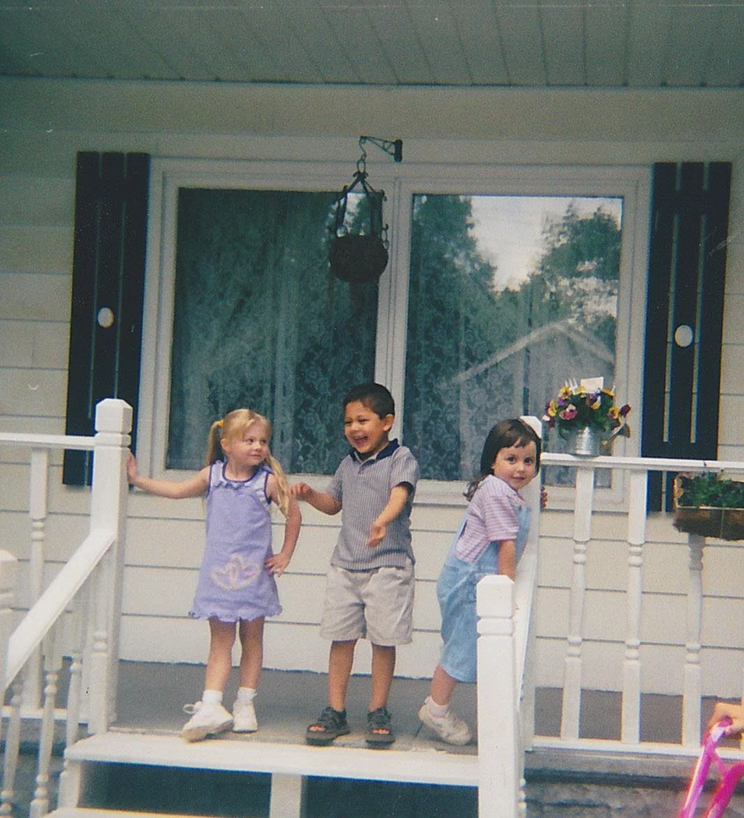 Three children, two girls and one boy, are standing on the porch of a house, holding hands and smiling. The girl on the left is wearing a purple dress with a swan design, the boy in the middle is wearing a striped shirt, and the girl on the right is in overalls. There are potted flowers on the porch railing, and a large window with a view of trees in the background.