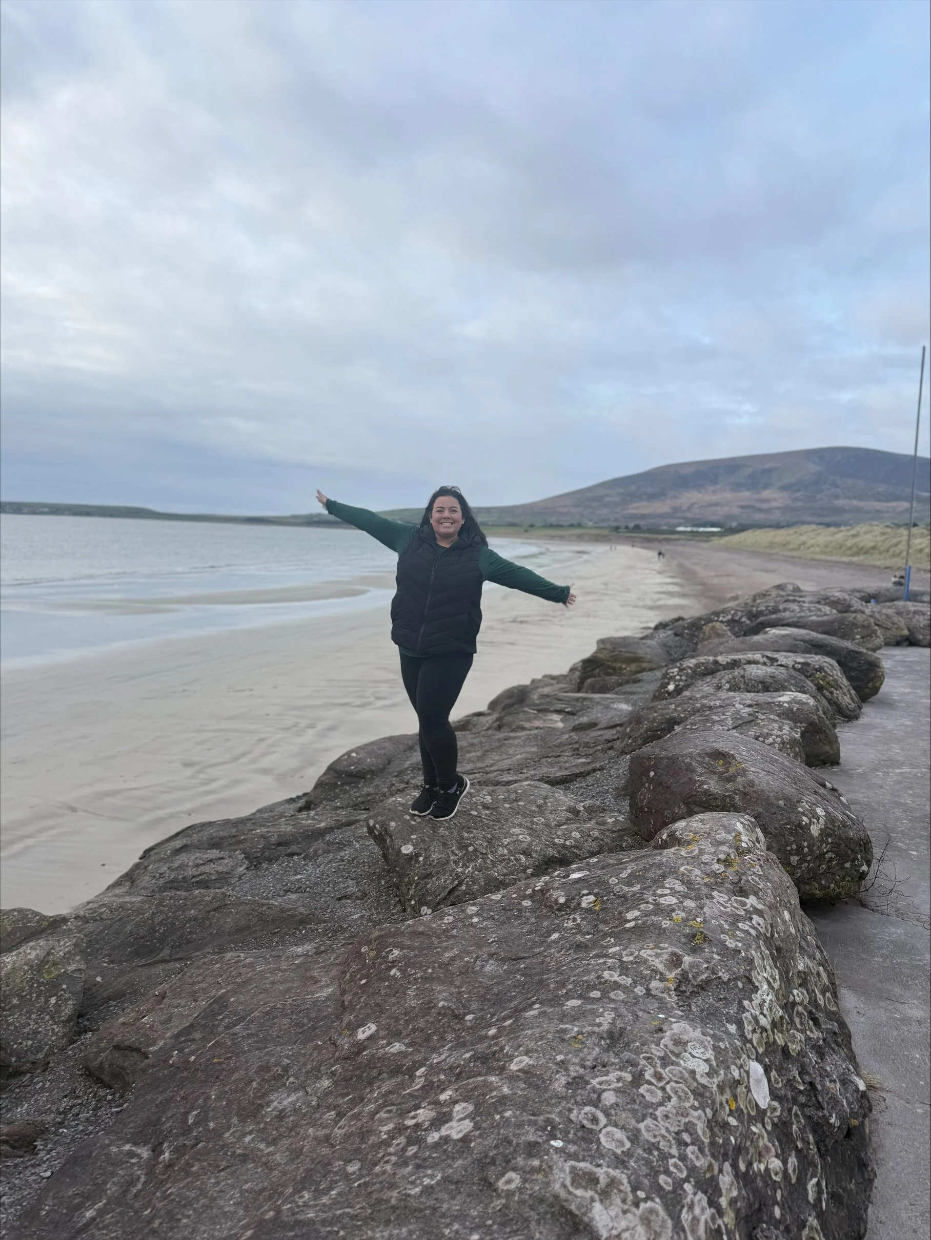 A smiling woman standing on large rocks at the edge of the beach with arms wide open, wearing a black puffer vest and black pants, with a body of water and a mountain in the background, under a cloudy sky.