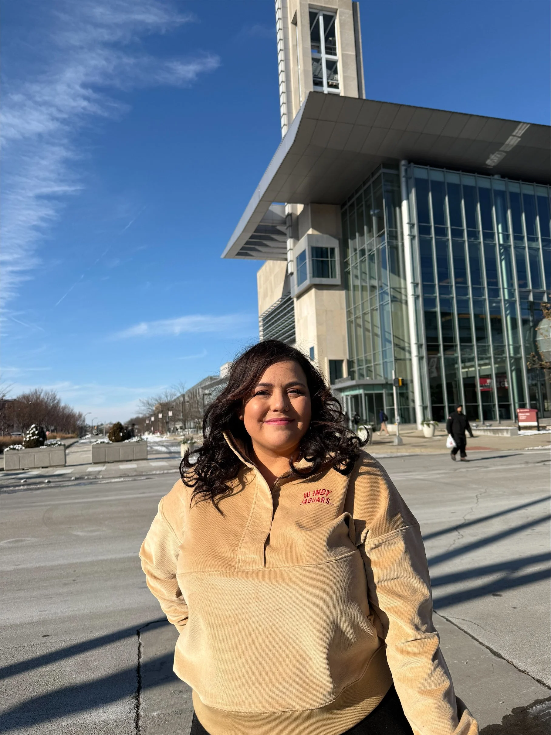 Young woman with dark curly hair smiling in front of a modern glass building on a sunny day.