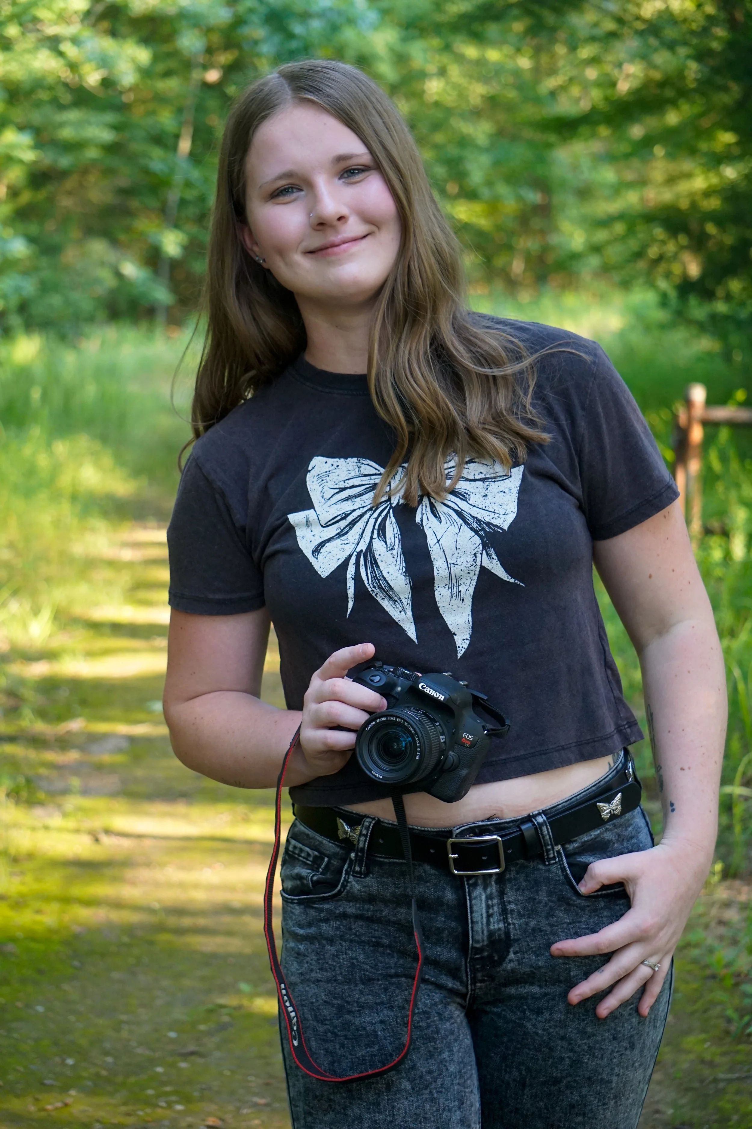 A young woman standing outdoors on a nature trail, holding a Canon DSLR camera, smiling at the camera, with green trees and grass in the background.