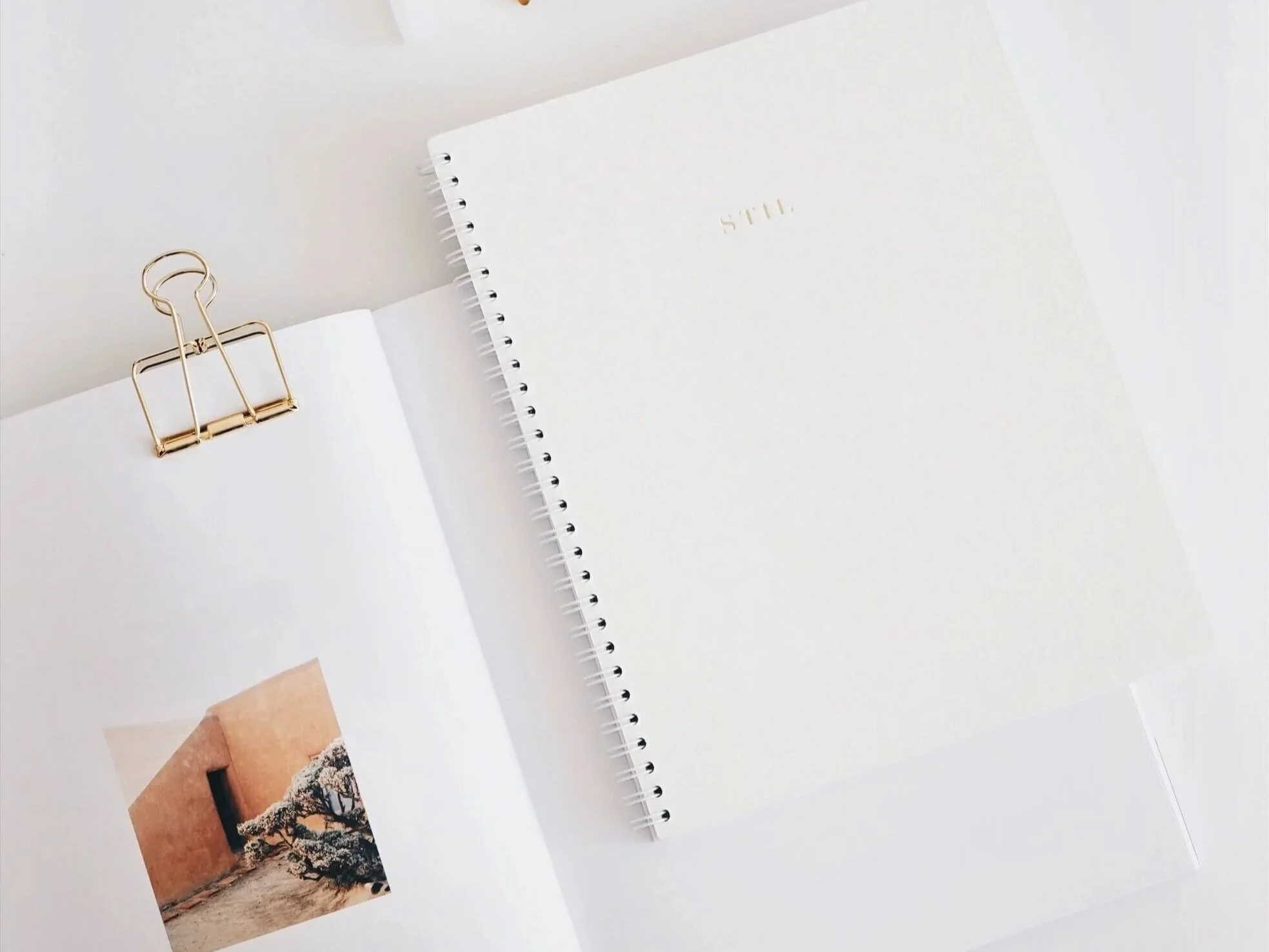 An open white notebook with a photo of desert landscape and a black tree inside, held by a gold binder clip, next to a closed white spiral notebook with "STILL" embossed on the cover, all on a white surface.