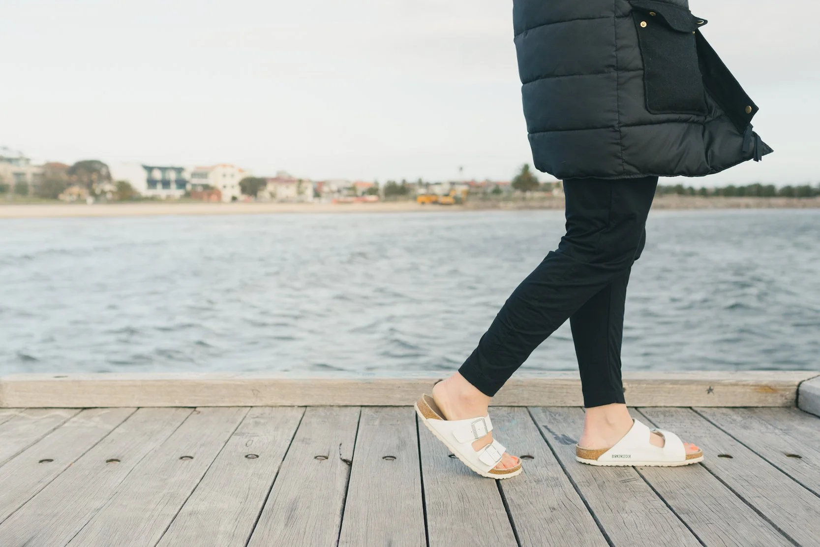 Person walking on a wooden pier next to a body of water, wearing black pants, white Birkenstock sandals, and a black puffer jacket, with buildings visible across the water.