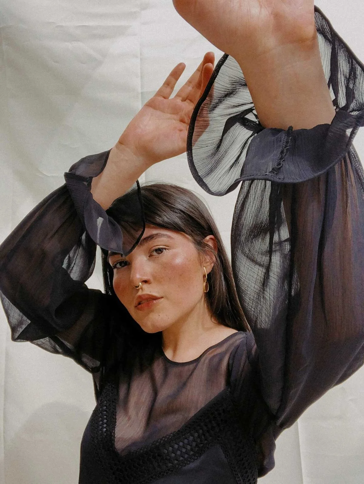 A woman with short dark hair wearing a sheer black top with puffed sleeves, gold hoop earrings, and a septum piercing, posing with her arms raised against a plain background.