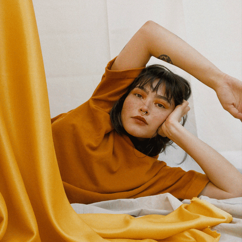 A young woman lying on a bed, resting her head on her left hand, with her right arm raised above her head. She is wearing an orange-brown shirt and has dark hair with bangs. The background is minimal and she is surrounded by yellow fabric.