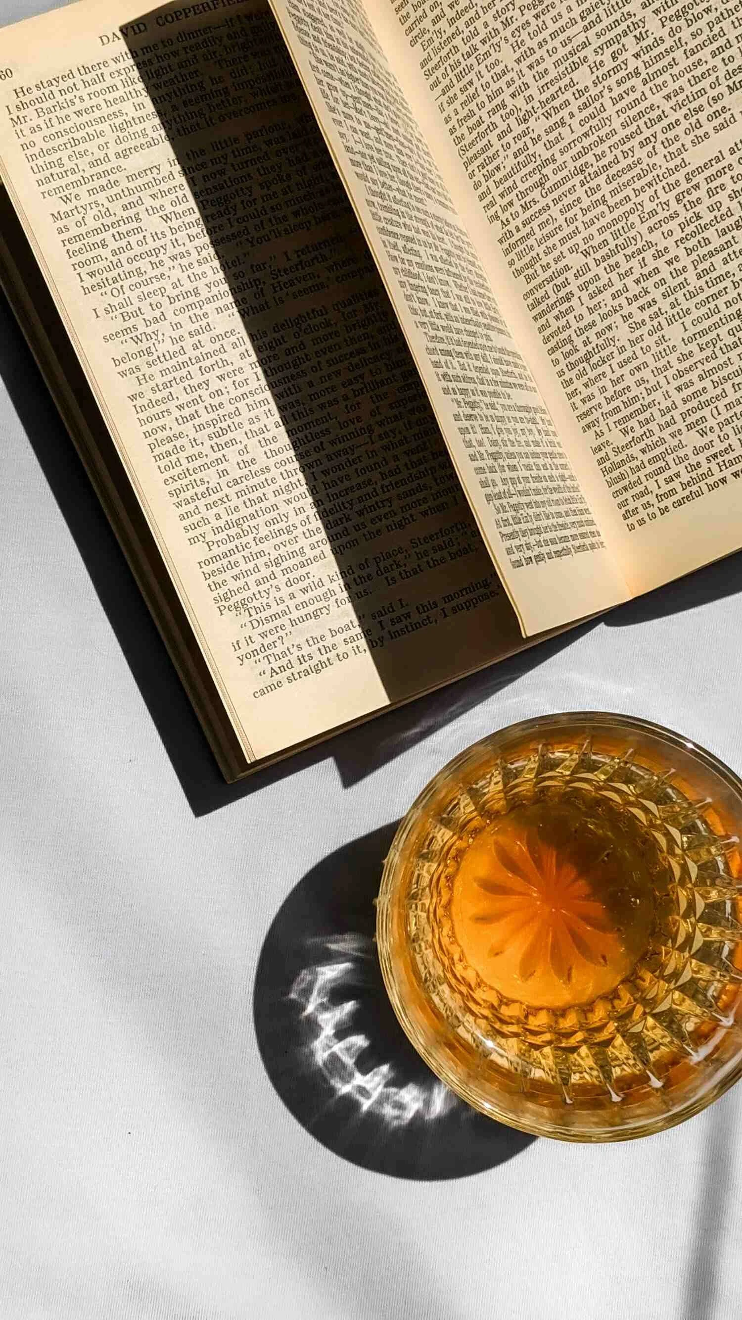 An open book with text and a glass of amber-colored beverage on a white surface. The beverage creates a decorative shadow on the surface.