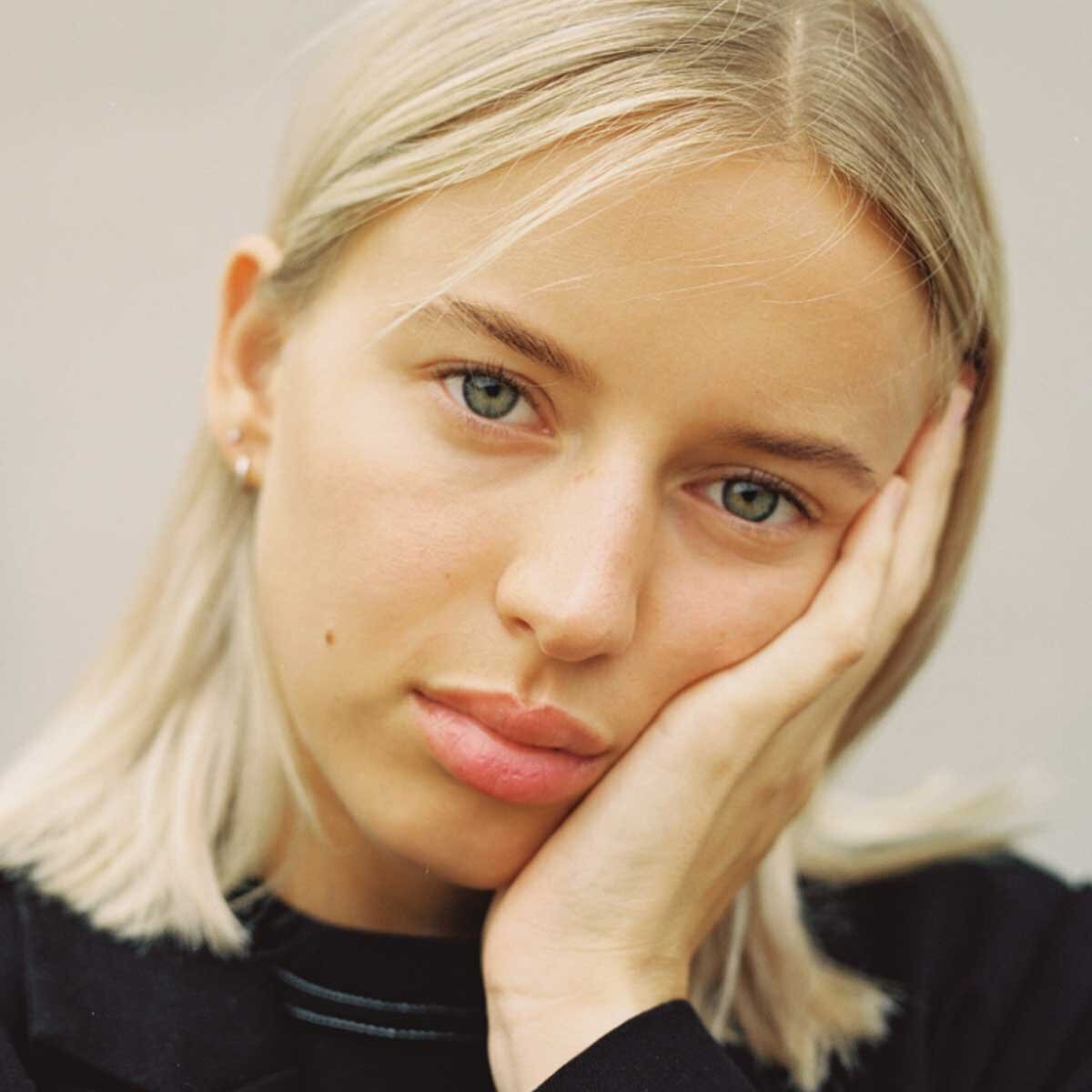 Close-up portrait of a young woman with blonde hair, blue eyes, and natural makeup, resting her face on her hand, wearing a black top.