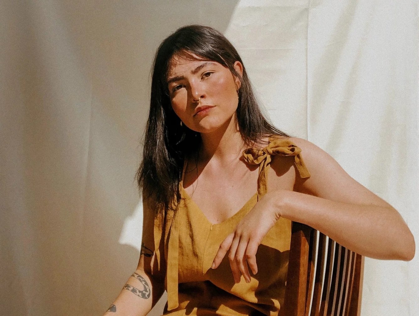 A woman with medium-length dark hair sitting on a wooden chair, wearing a tan sleeveless top with tied shoulder straps, posing with her arm resting on the back of the chair, against a plain wall background.