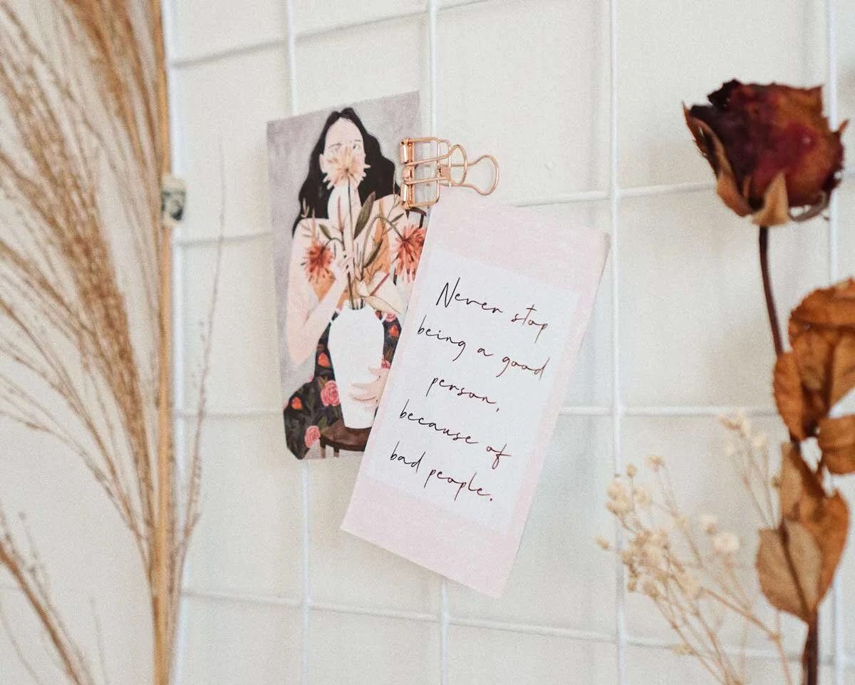 A decorative wall with dried flowers, an illustrated woman holding a plant, and a handwritten note that reads, 'Never stop being a good person, because of bad people.'