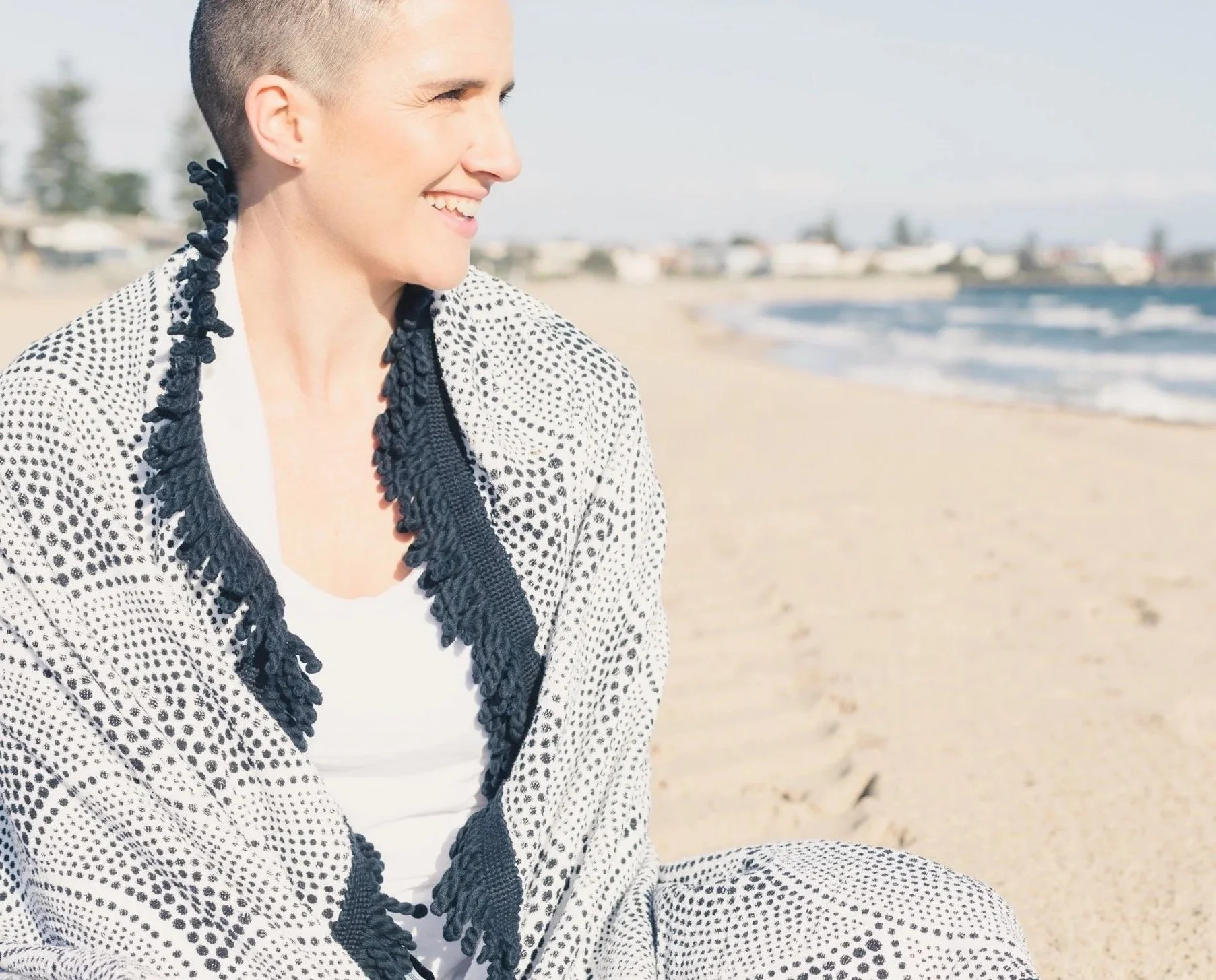 A woman sitting on a sandy beach, smiling and looking to her left, with the ocean and distant buildings in the background.