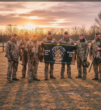Seven hunters in camouflage clothing standing outdoors at sunset, holding a banner that reads 'Mettowee Outdoor' and mentions 'United We Drive, Divided We Fall'.