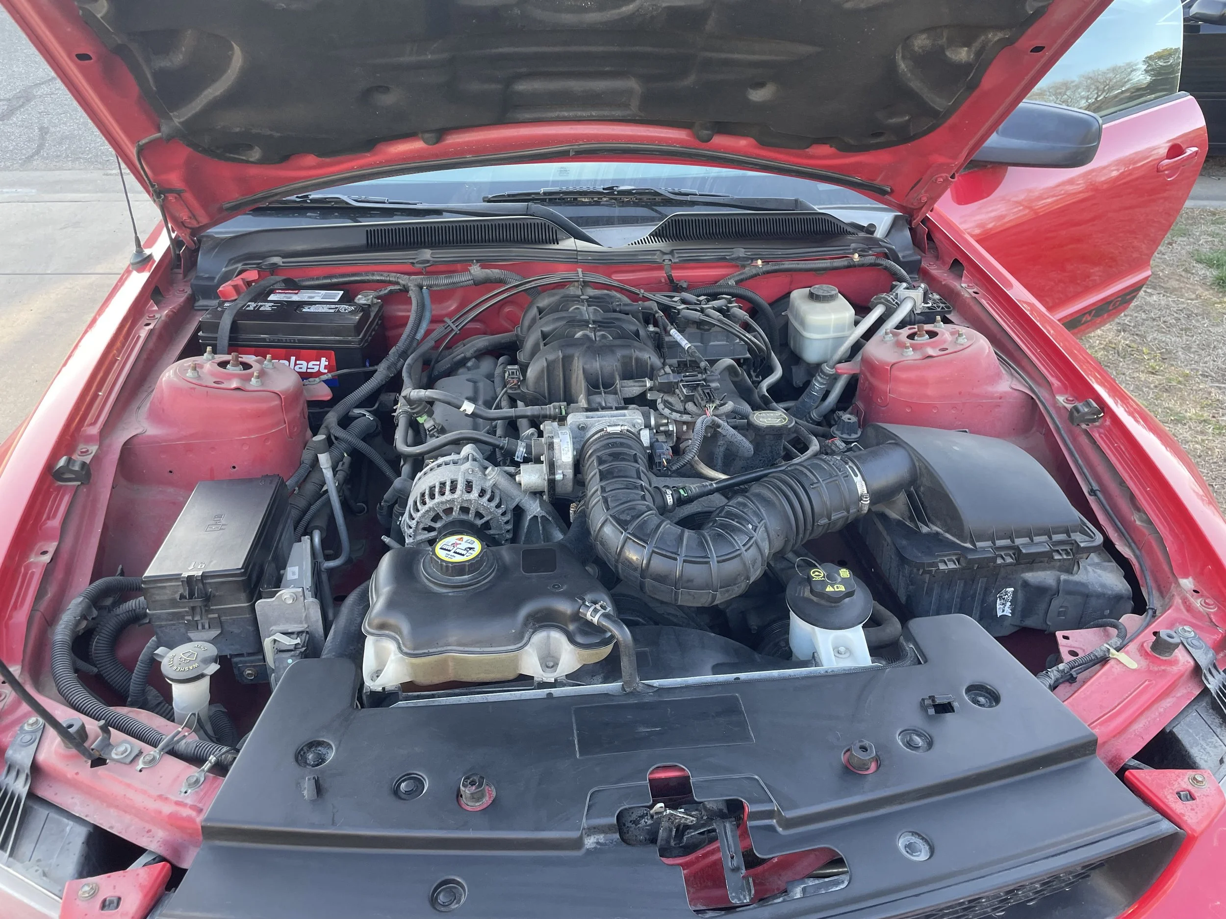 The engine bay of a red car showing various components including the battery, air intake, alternator, and coolant reservoir.