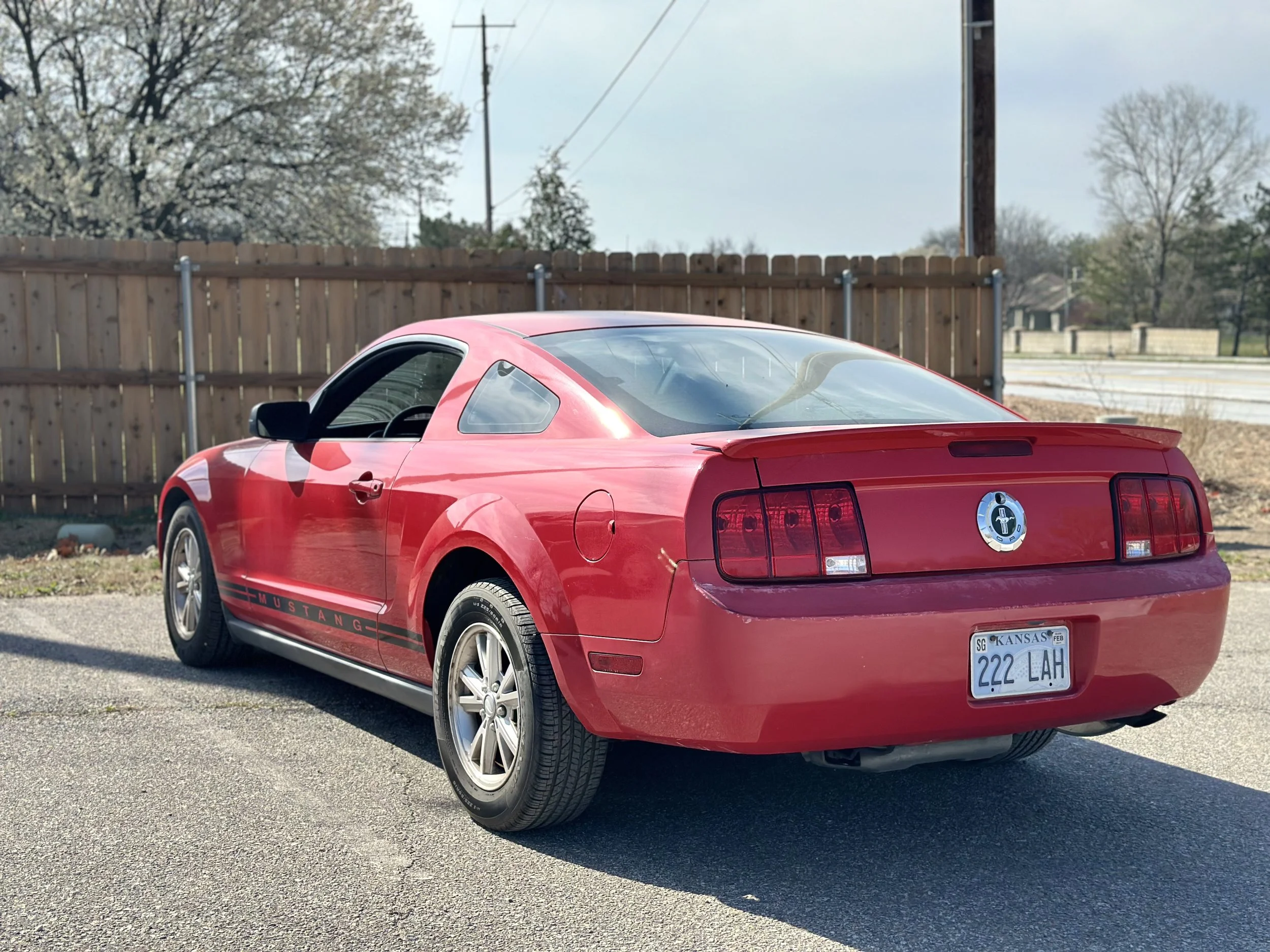 Red Ford Mustang with Kansas license plate 222 LAH parked on street near a wooden fence.
