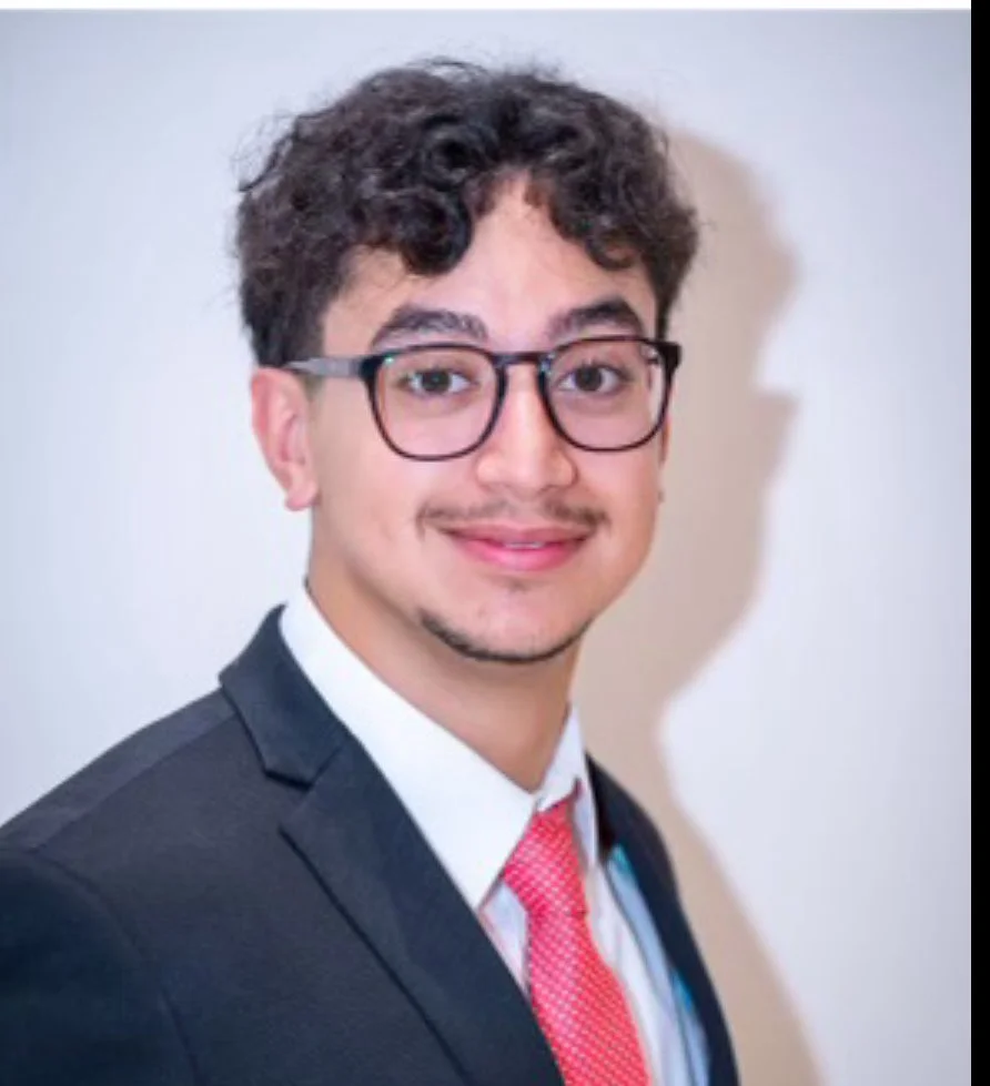 A young man wearing glasses, a dark suit, white shirt, and a red patterned tie, smiling confidently against a plain white background.