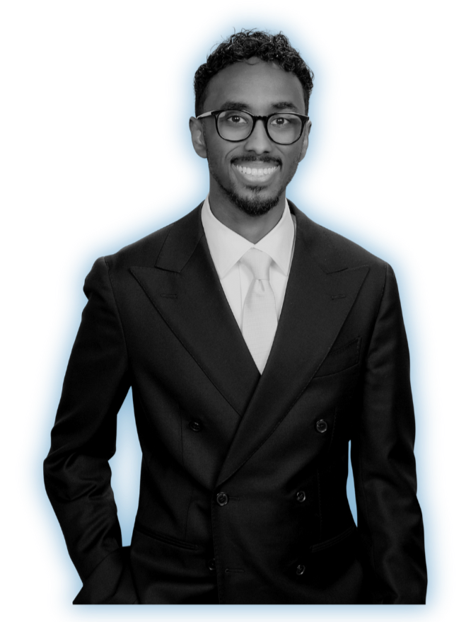 A man wearing glasses in a black double-breasted suit, white shirt, and tie, smiling and standing with one hand in his pocket.