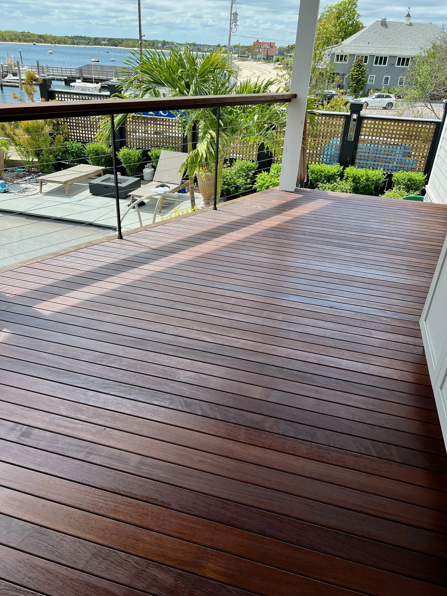 A wooden deck overlooking a waterfront scene with boats, a pier, and residential houses, surrounded by greenery and plants.