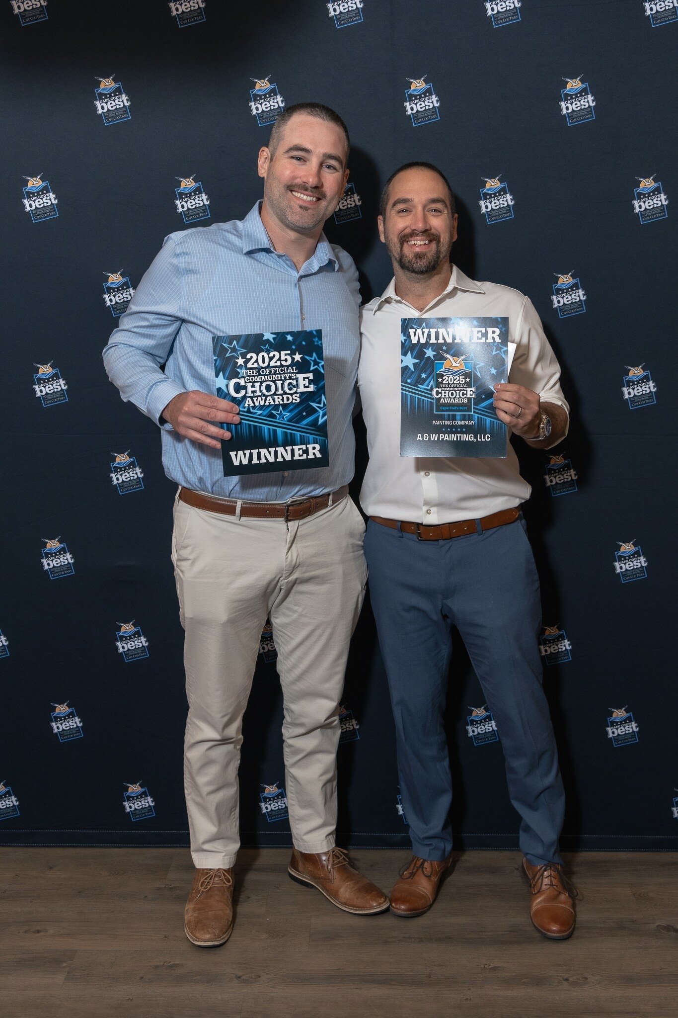 Two men standing in front of a backdrop with a logo pattern, holding awards that read "2025 The Official Community Choice Awards Winner". The man on the left wears a light blue shirt and beige pants, while the man on the right wears a white shirt and blue pants.