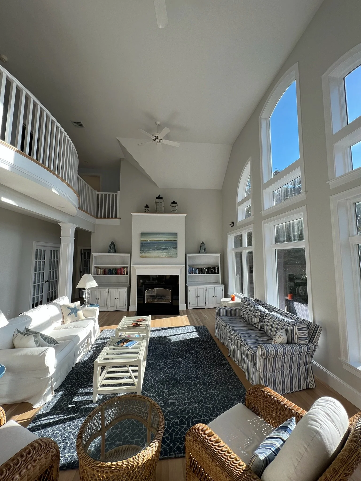 Living room with large windows, white furniture, striped and wicker chairs, bookshelf, fireplace, and nautical decor.