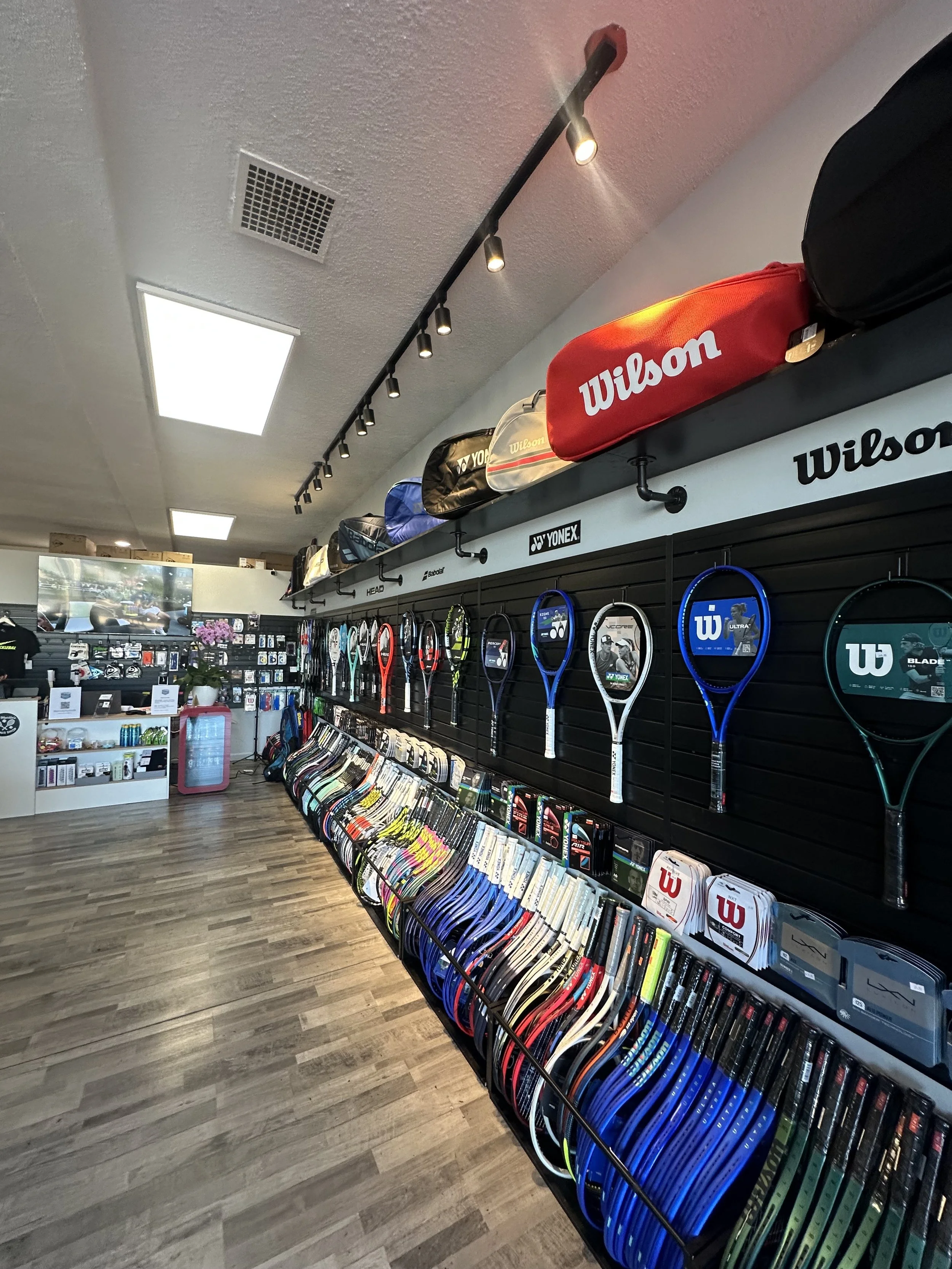 A sports store displays tennis rackets, tennis bags, and accessories on black slatwall panels and shelves, with a TV and checkout counter in the background.