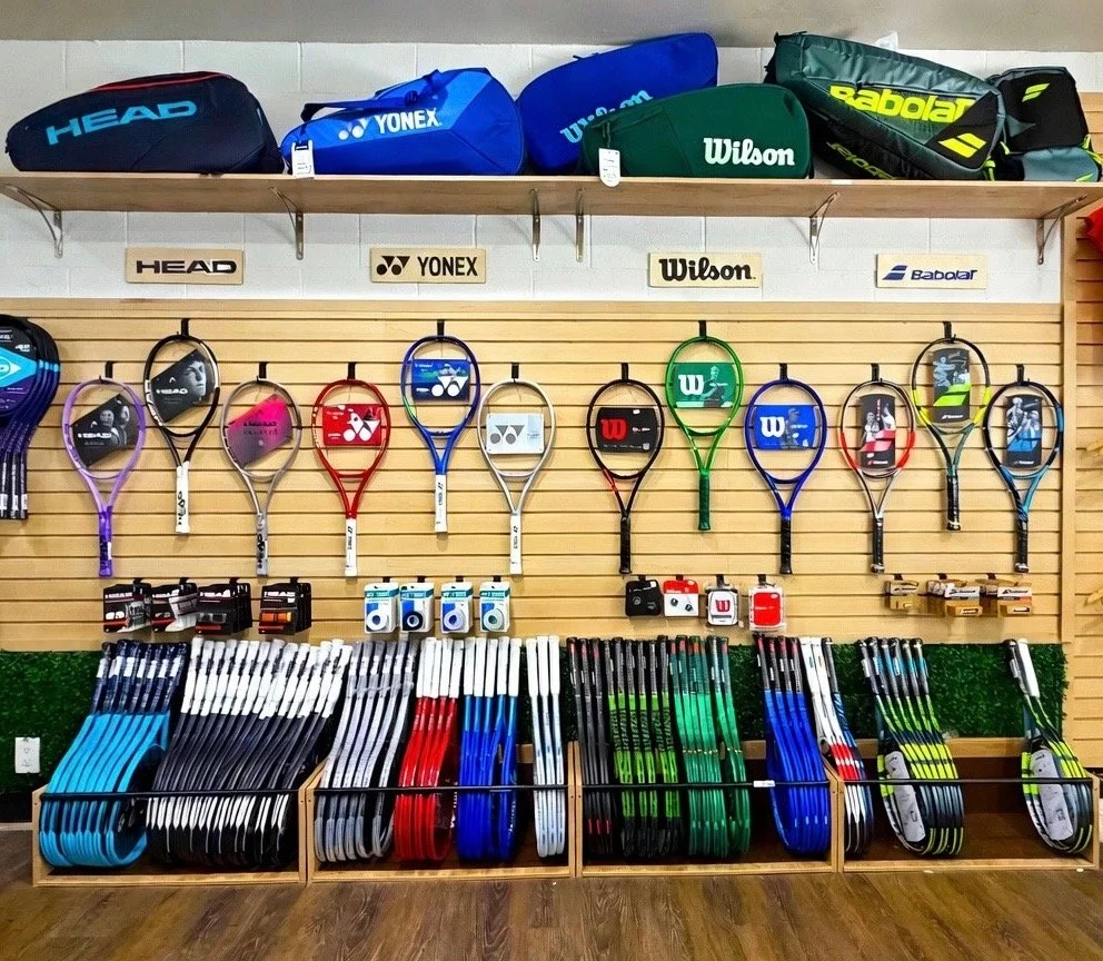 A display of tennis rackets and accessories in a sports store. The rackets are arranged on a wooden slat wall, with some stacked in a row at the bottom. Various brands of tennis bags are on a shelf above the rackets, including HEAD, YONEX, Wilson, and Babolat.