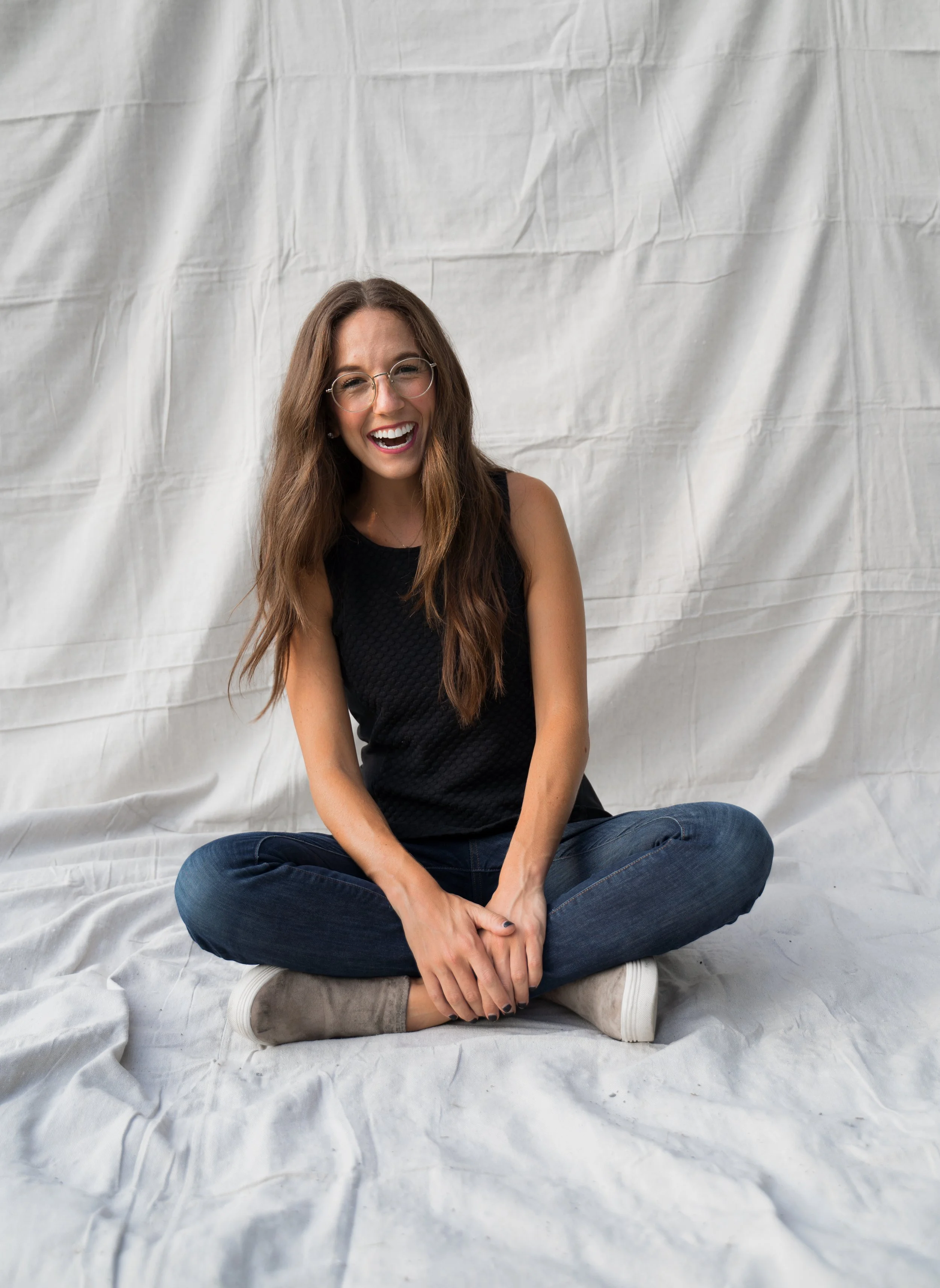 A woman sitting cross-legged on a white fabric background, smiling and laughing, wearing glasses, a black sleeveless top, blue jeans, and beige shoes.