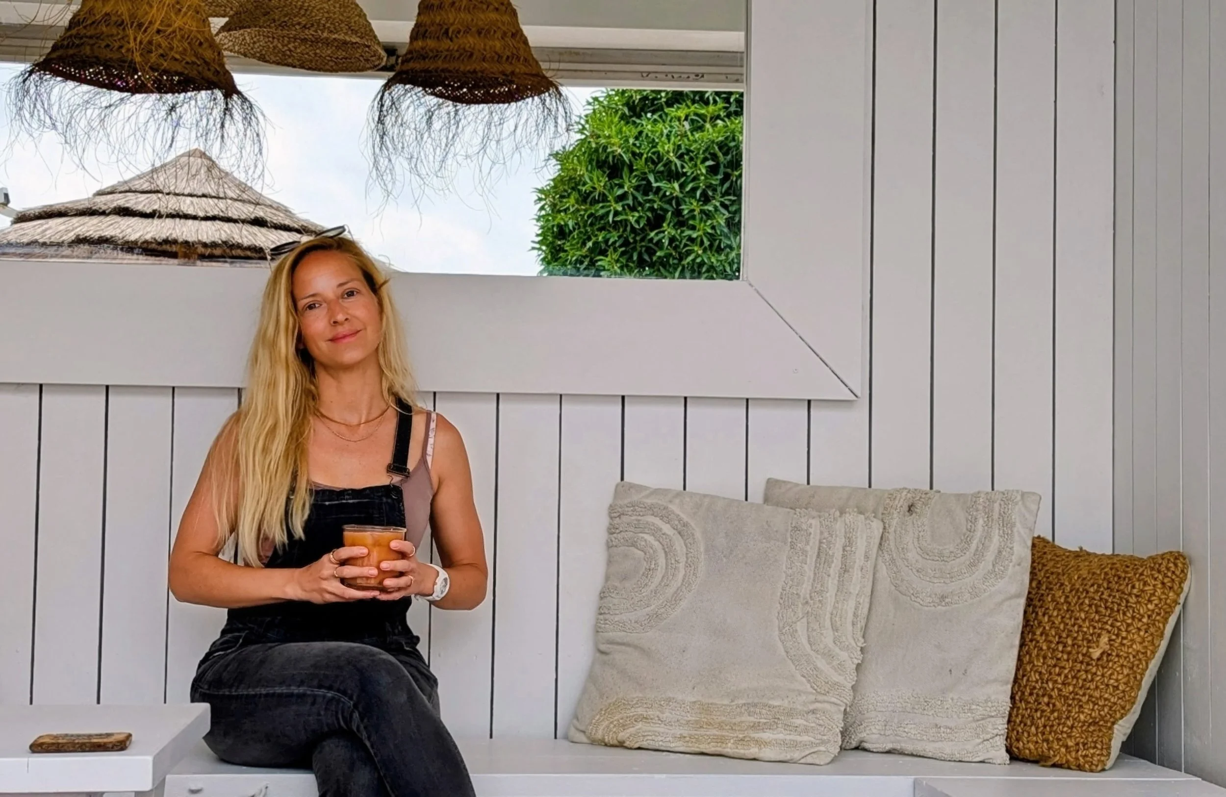 A woman with long blonde hair sitting on a white wooden bench, holding a glass of drink, with pillows behind her and a window showing a thatched umbrella and green trees outside.