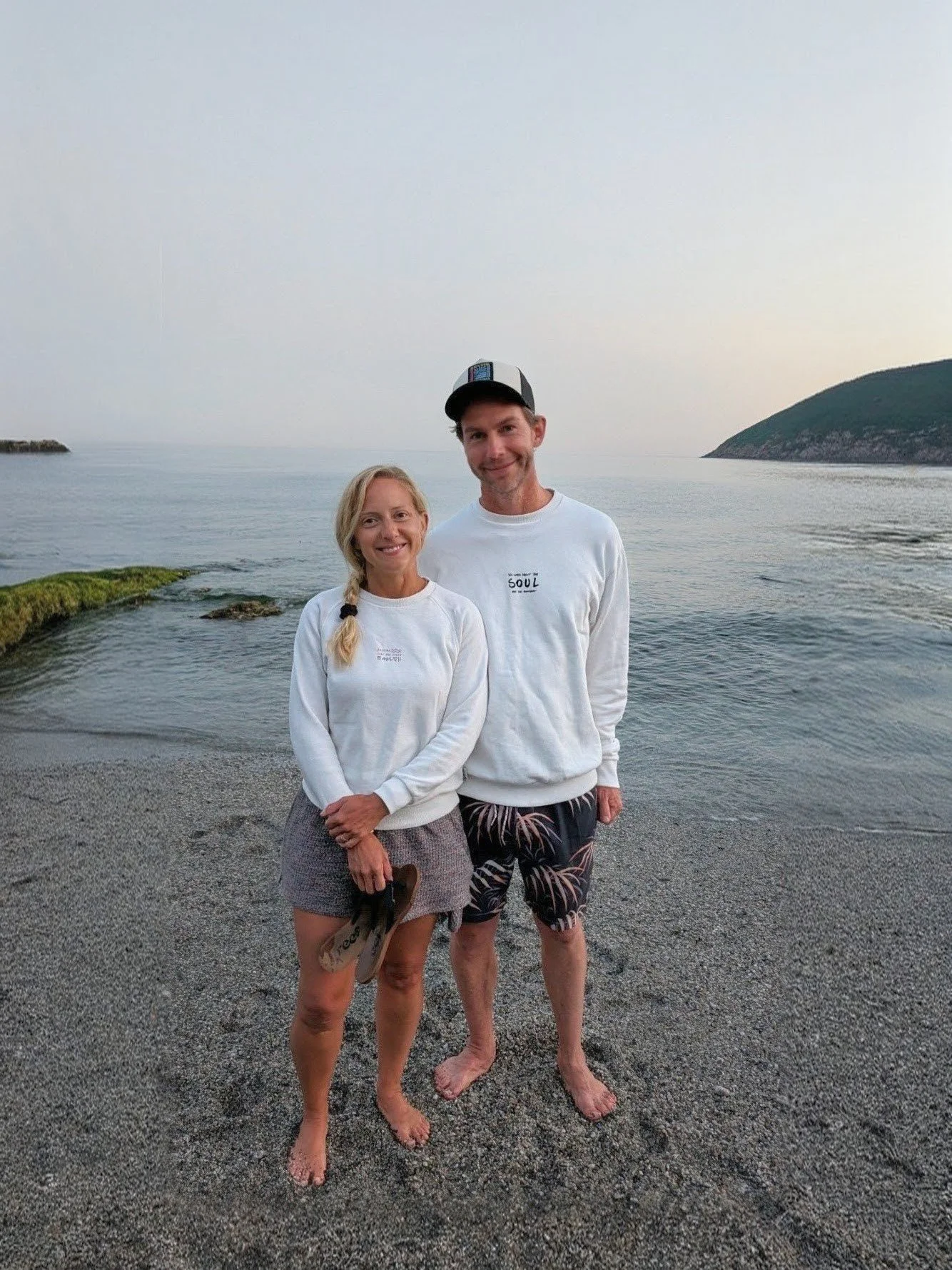 A smiling woman and man standing on a pebble beach near the ocean, with a small hill or mountain in the background. Both are wearing white sweatshirts, and the woman is holding a pair of flip-flops. It is dusk or early evening.