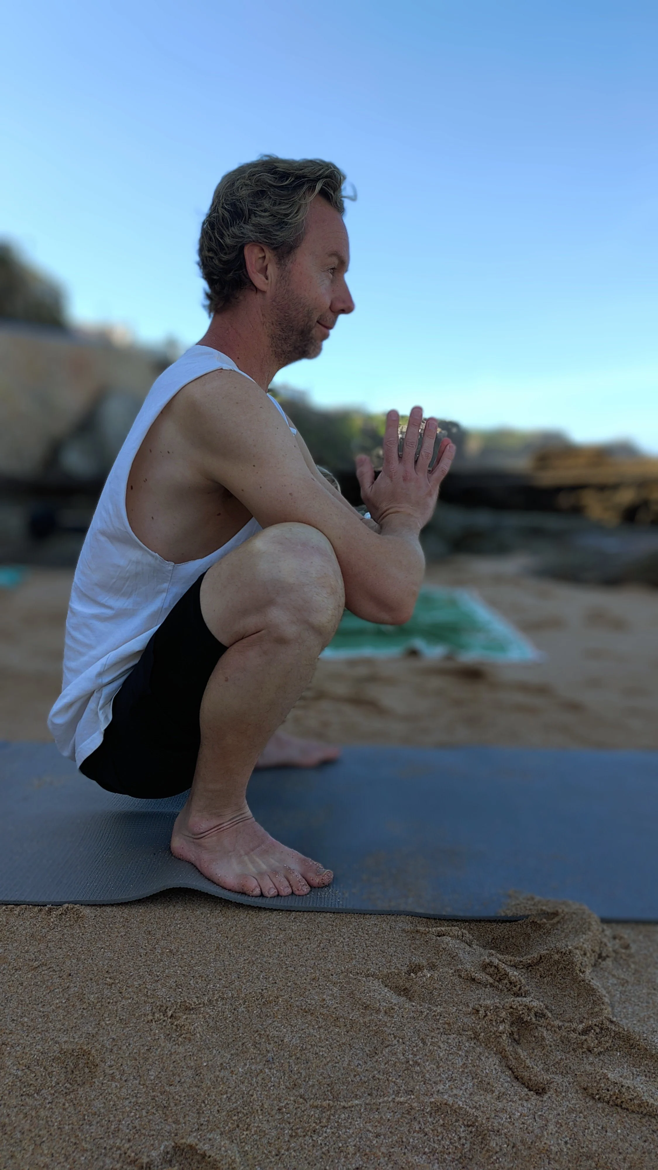 A man practicing yoga on a beach, squatting on a yoga mat with hands pressed together in a prayer position, looking peaceful.