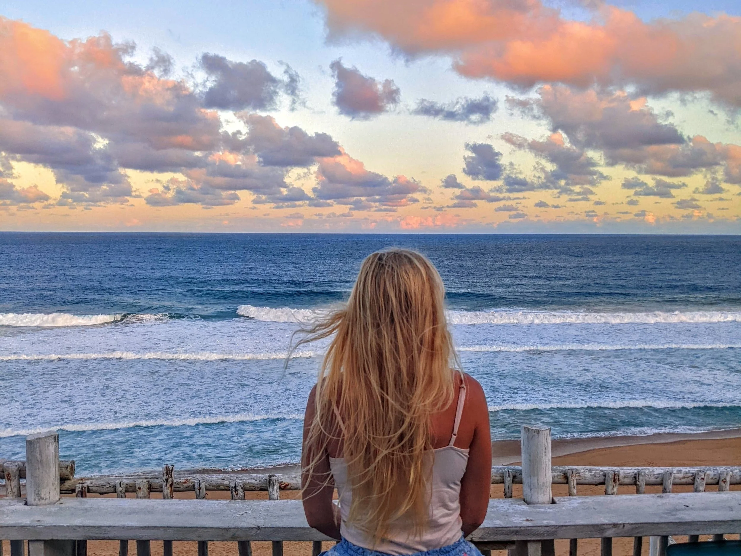 A woman with long blonde hair sitting on a wooden bench, facing the ocean with waves and a colorful sky with clouds during sunset.
