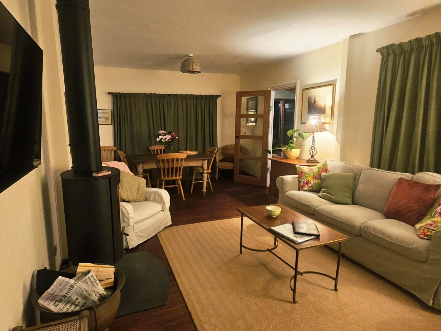 A cozy living room with a beige sofa and a smaller matching loveseat, decorated with colorful pillows. A wooden coffee table with magazines and a bowl sits on a beige rug, with green curtains on the windows. In the background, a dining area with a wo