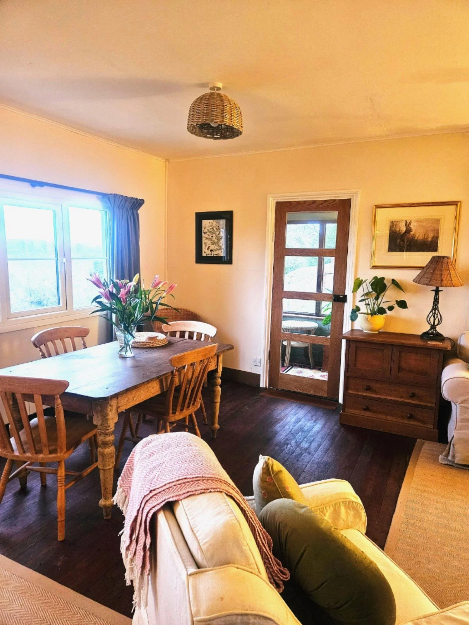 A cozy living room with a wooden dining table, lots of chairs, a cream sofa with a pink blanket, a large window with blue curtains, a wooden sideboard with a yellow plant pot and a lamp, artwork on the walls, and a door with glass panels leading to another room.