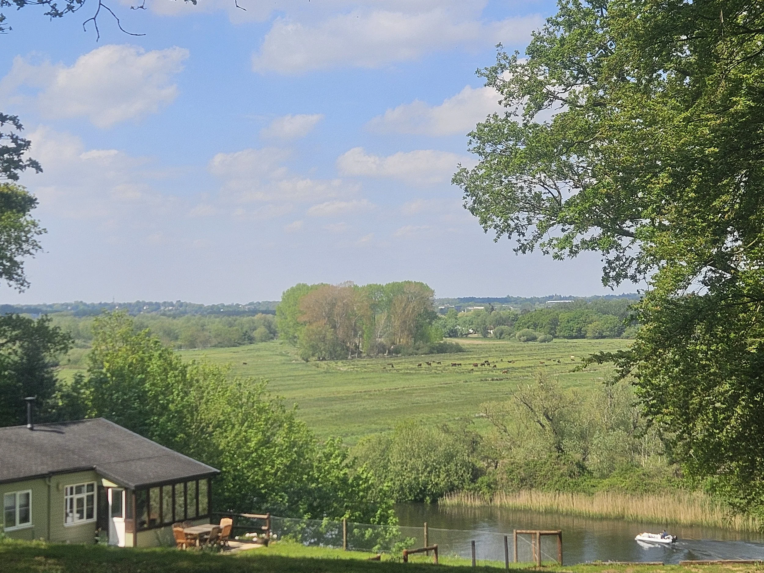 Scenic view of a river with trees, green fields, and a house in the foreground, with a bright blue sky and scattered clouds.