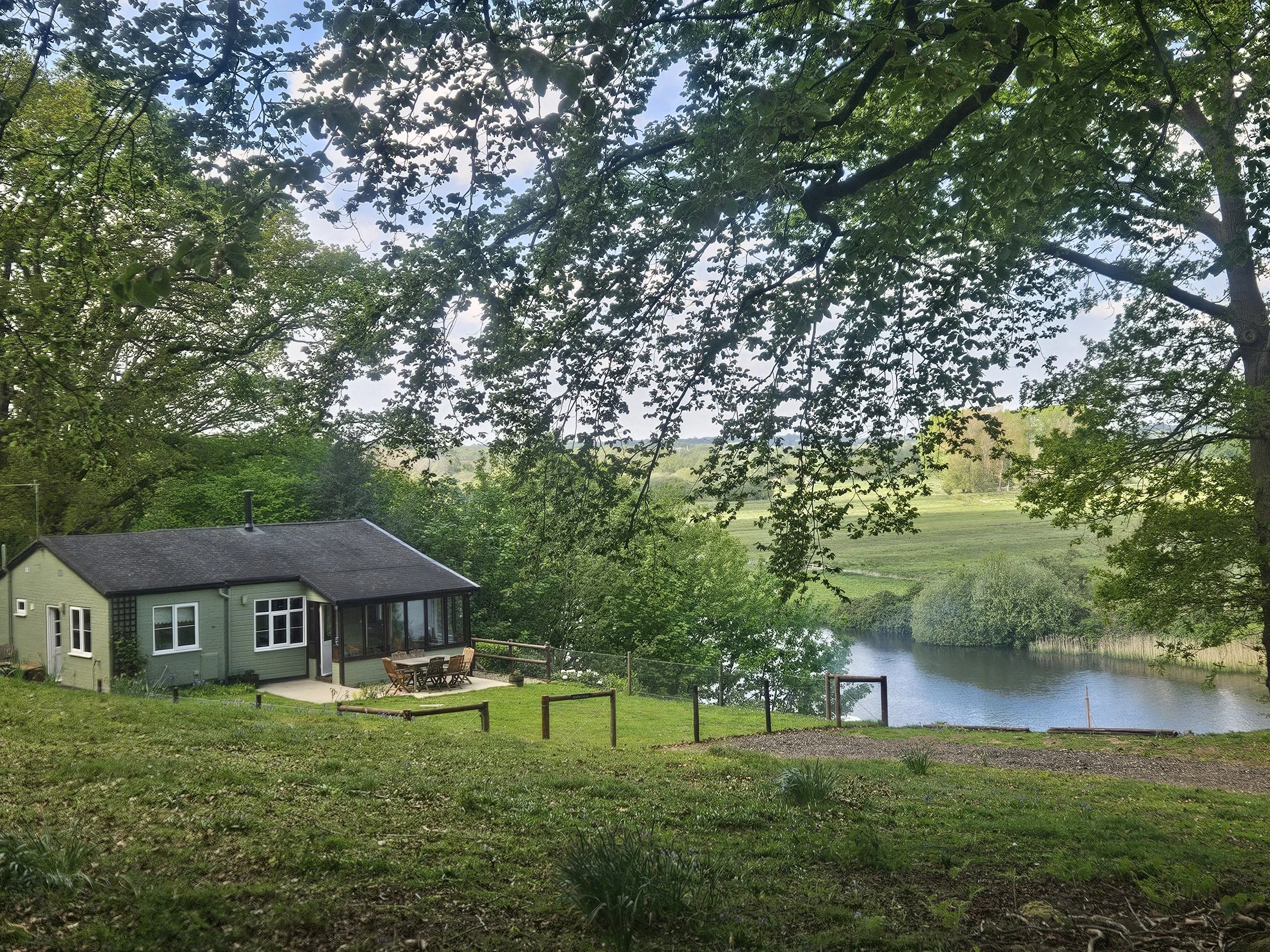 A lakeside backyard scene with a house featuring large windows and a patio with dining furniture, surrounded by green trees and grass, with a body of water in the background.