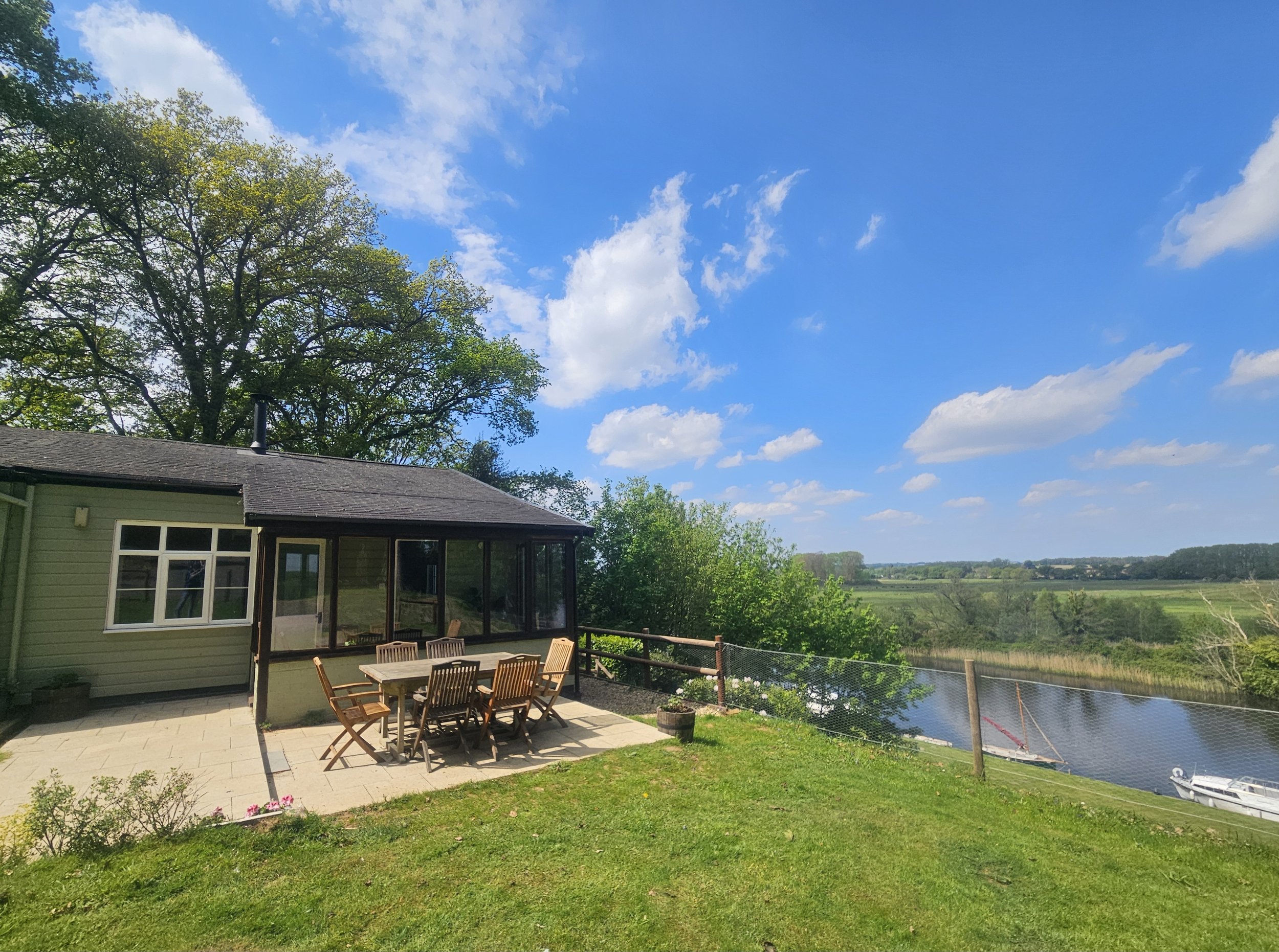 A backyard patio with a wooden dining table and chairs, a screened porch, green lawn, and a body of water with a boat dock, surrounded by trees and open fields under a partly cloudy blue sky.