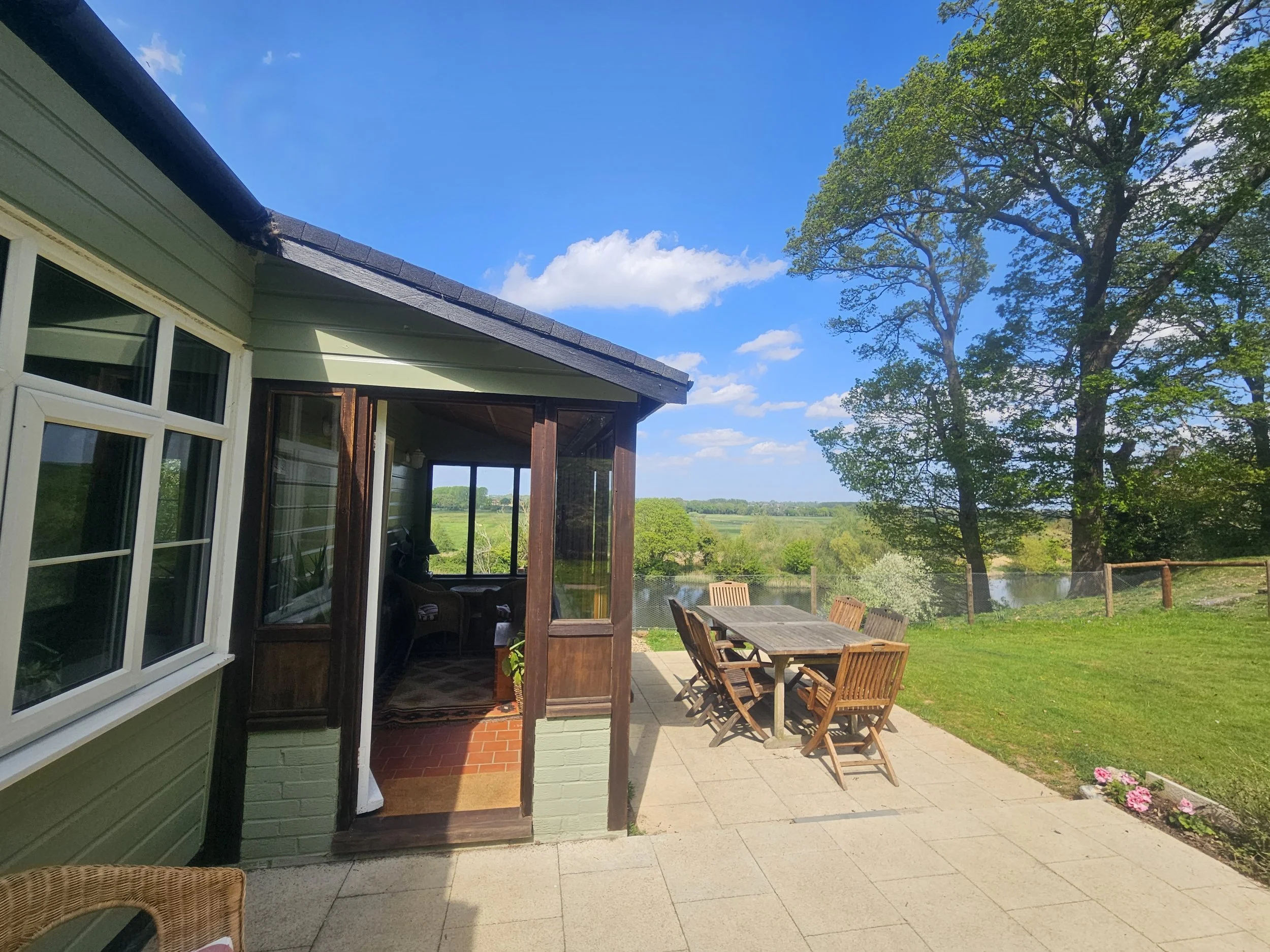 View of a house's back patio area with a wooden dining table and chairs, surrounded by a green lawn, trees, and a river in the distance under a blue sky with some clouds.