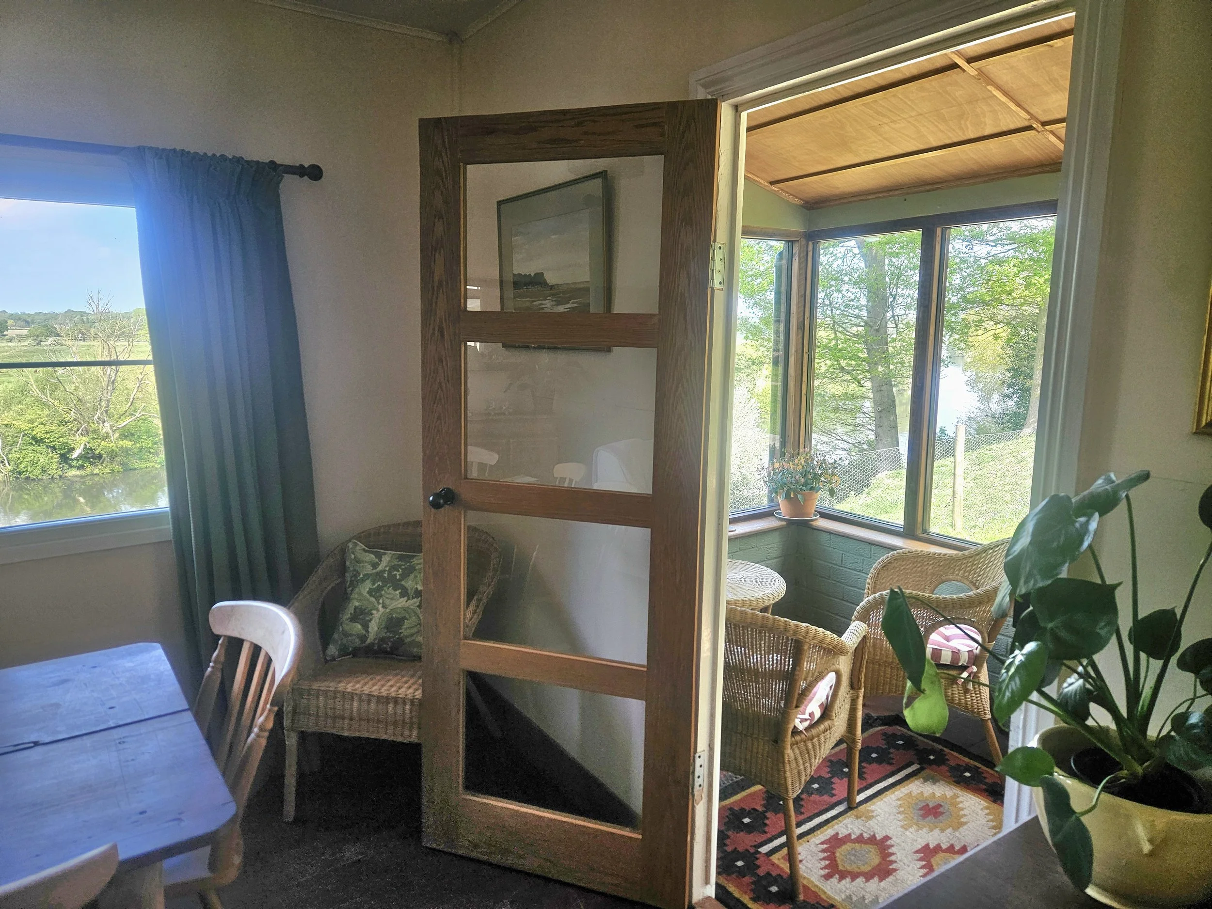 Interior of a house showing a room with a wooden cabinet mirror, outdoor view through windows, wicker chairs, colorful rug, and house plants.
