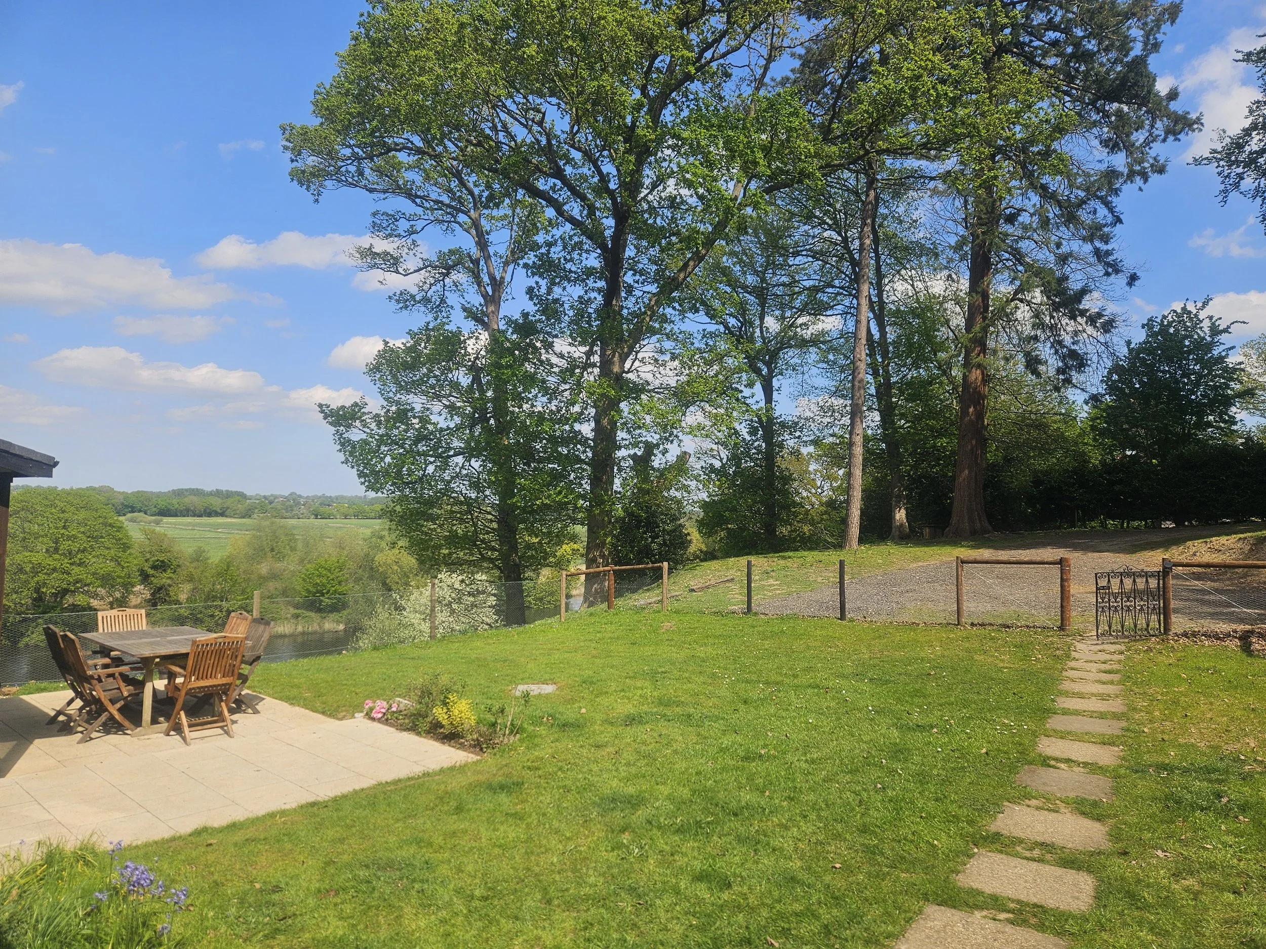 A backyard with a patio, wooden chairs and table, green lawn, blooming flowers, tall trees, and a bright blue sky with some clouds.