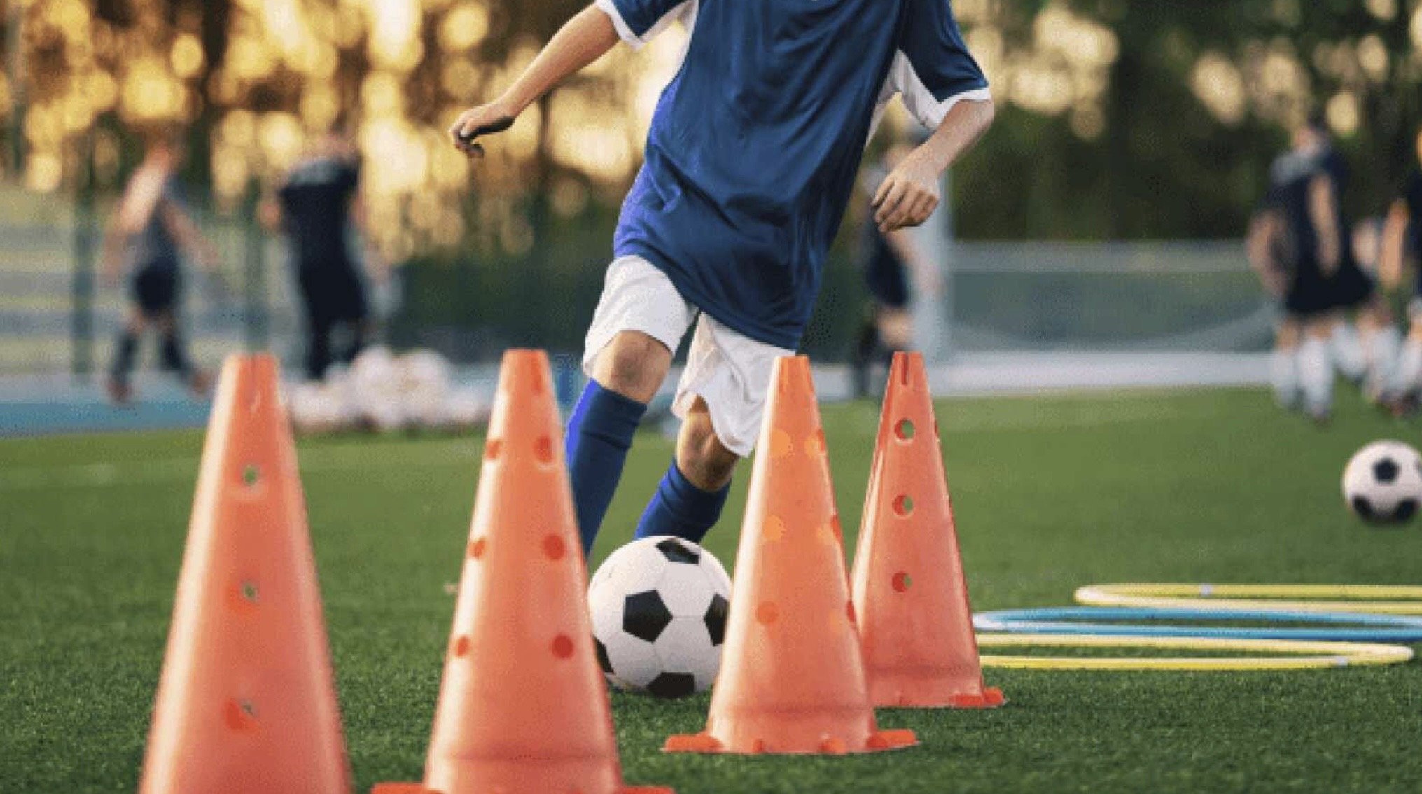 A young soccer player practicing dribbling skills with a soccer ball on a field, surrounded by orange training cones and agility rings, with teammates in the background.