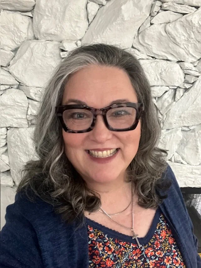 A woman with gray hair, wearing large tortoise-shell glasses, smiling, against a white stone wall.