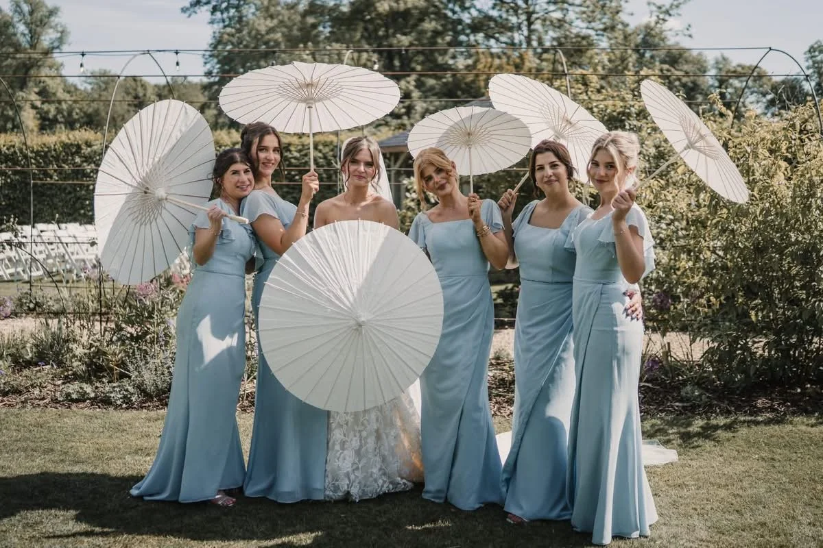 Group of six women in light blue dresses holding white parasols outdoors during a sunny day, standing on grass with trees and garden in the background.