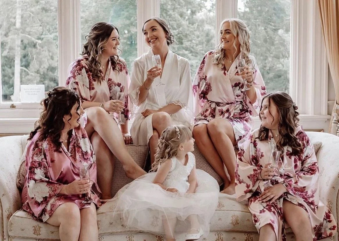 A group of six women and a young girl in various floral robes and a white dress, sitting and standing on a vintage sofa in a room with large windows and curtains, smiling and chatting, holding glasses of champagne.
