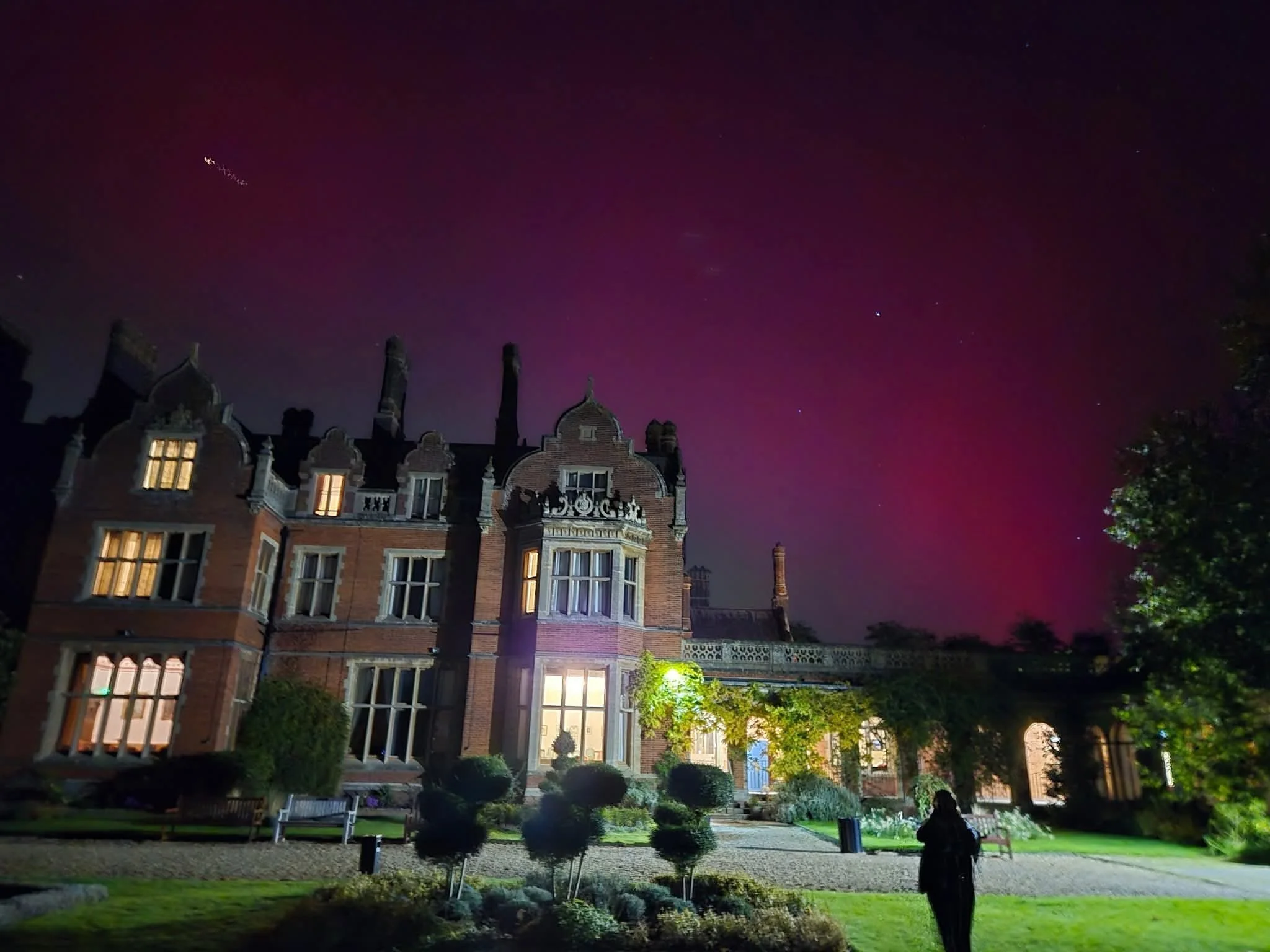 Night view of a large, historic brick mansion with illuminated windows, surrounded by landscaped gardens, under a starry sky with a hint of purple aurora.
