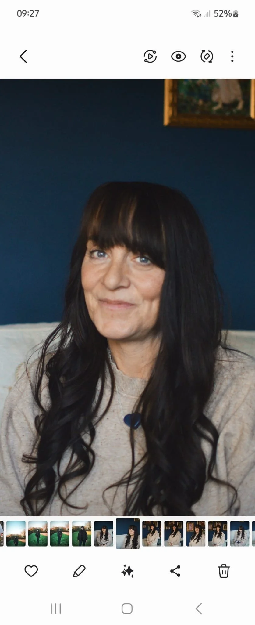 A woman with long, wavy black hair and bangs, wearing a beige top, sitting indoors against a dark blue wall.